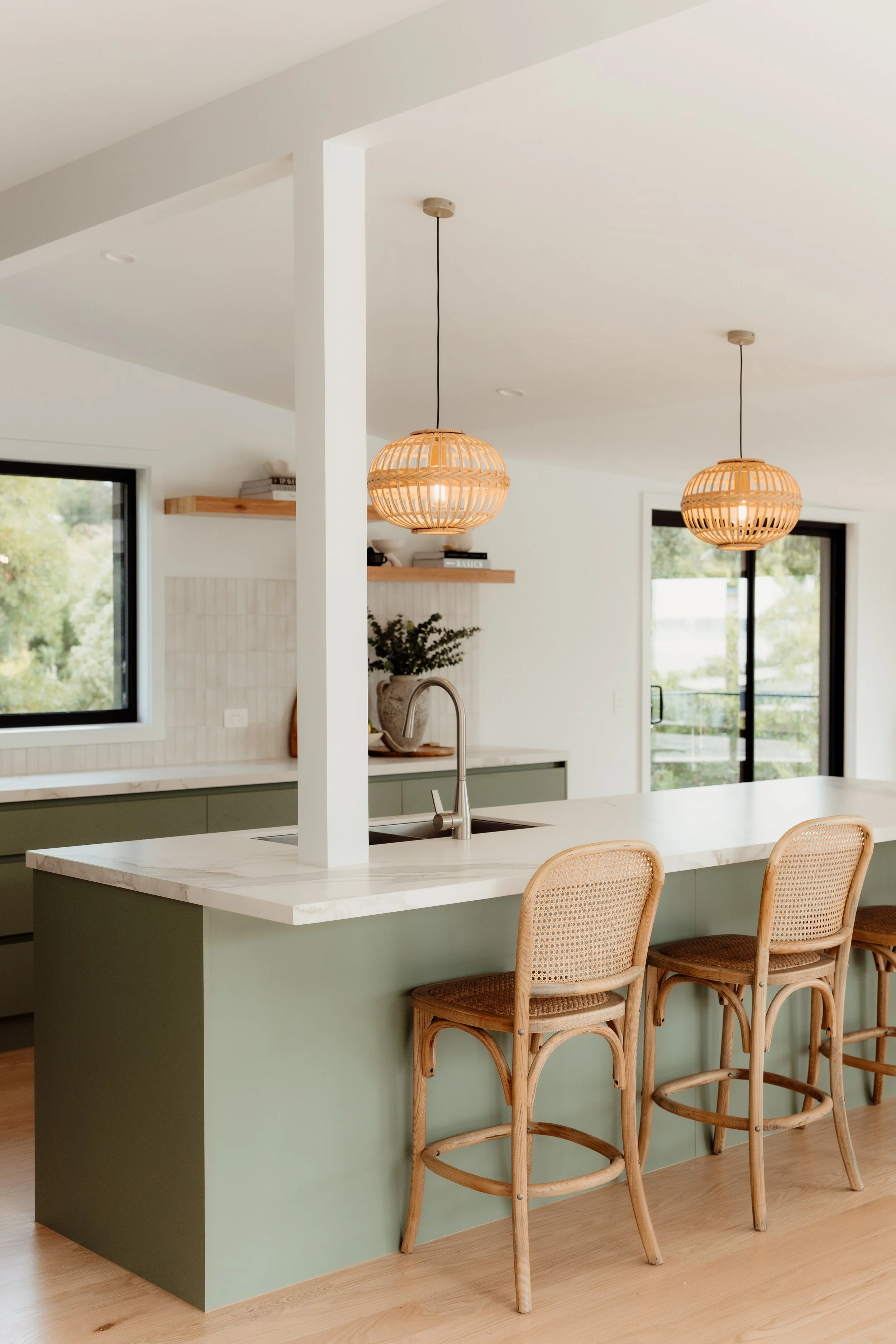 A modern kitchen with a white marble island, green base cabinet, and four woven rattan barstools. Two round wicker pendant lights hang above the island, and there are large windows and a sliding glass door to the outside.