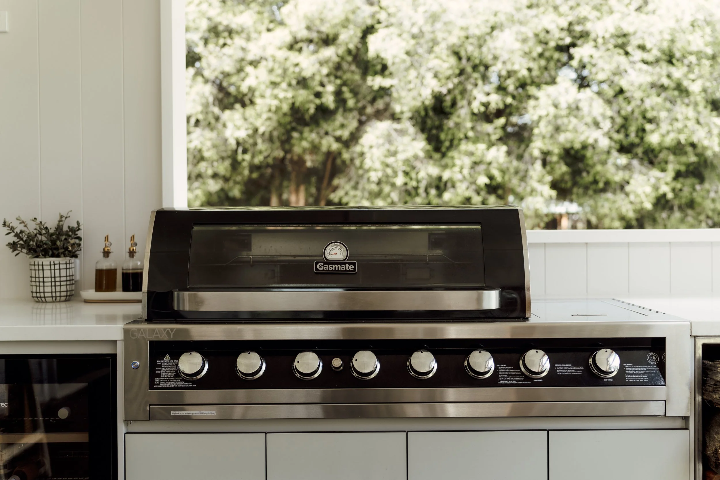 Outdoor gas grill with control knobs on a white kitchen counter, surrounded by potted plants and condiments, with a view of trees outside through a large window.