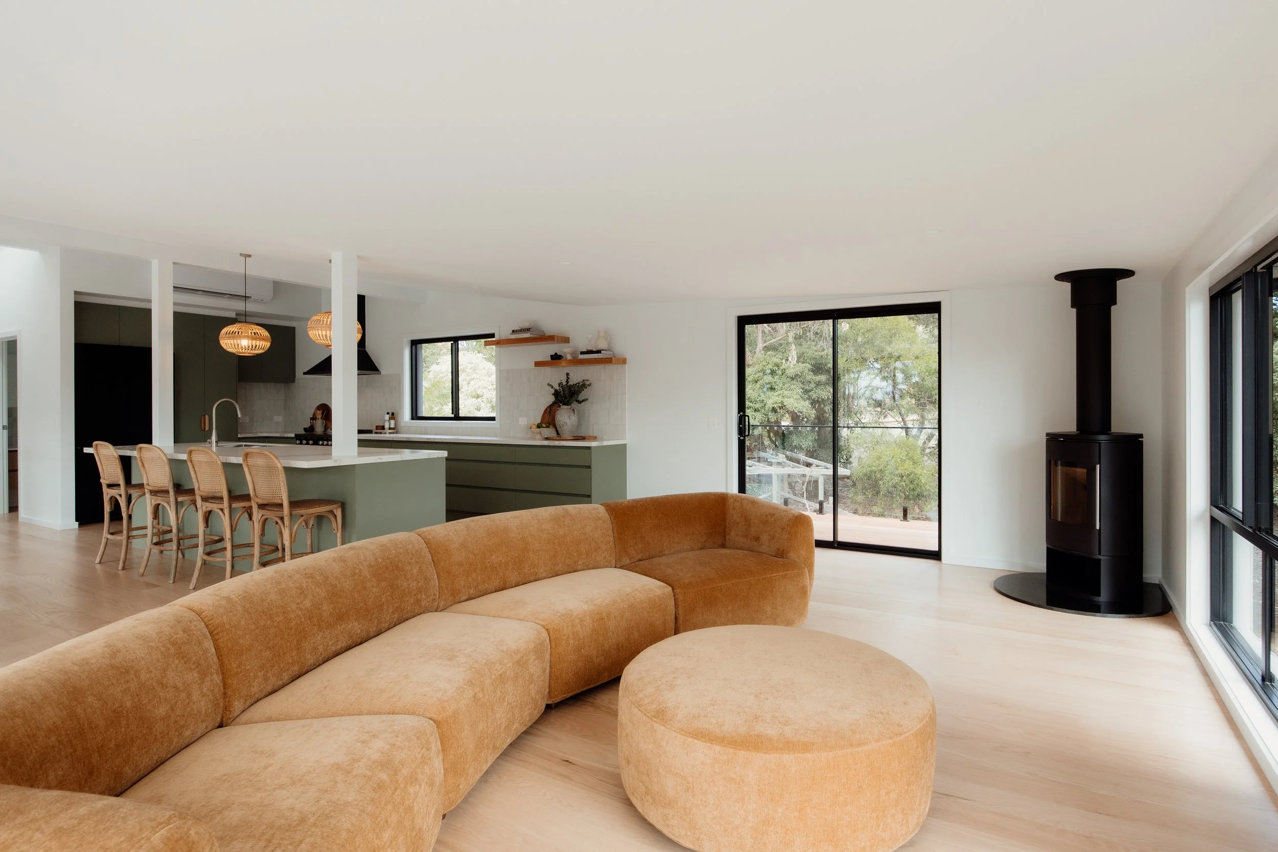 Modern open-concept living room and kitchen with beige sectional sofa, wooden dining chairs, kitchen island, hanging pendant lights, large windows, and sliding glass door leading outside.