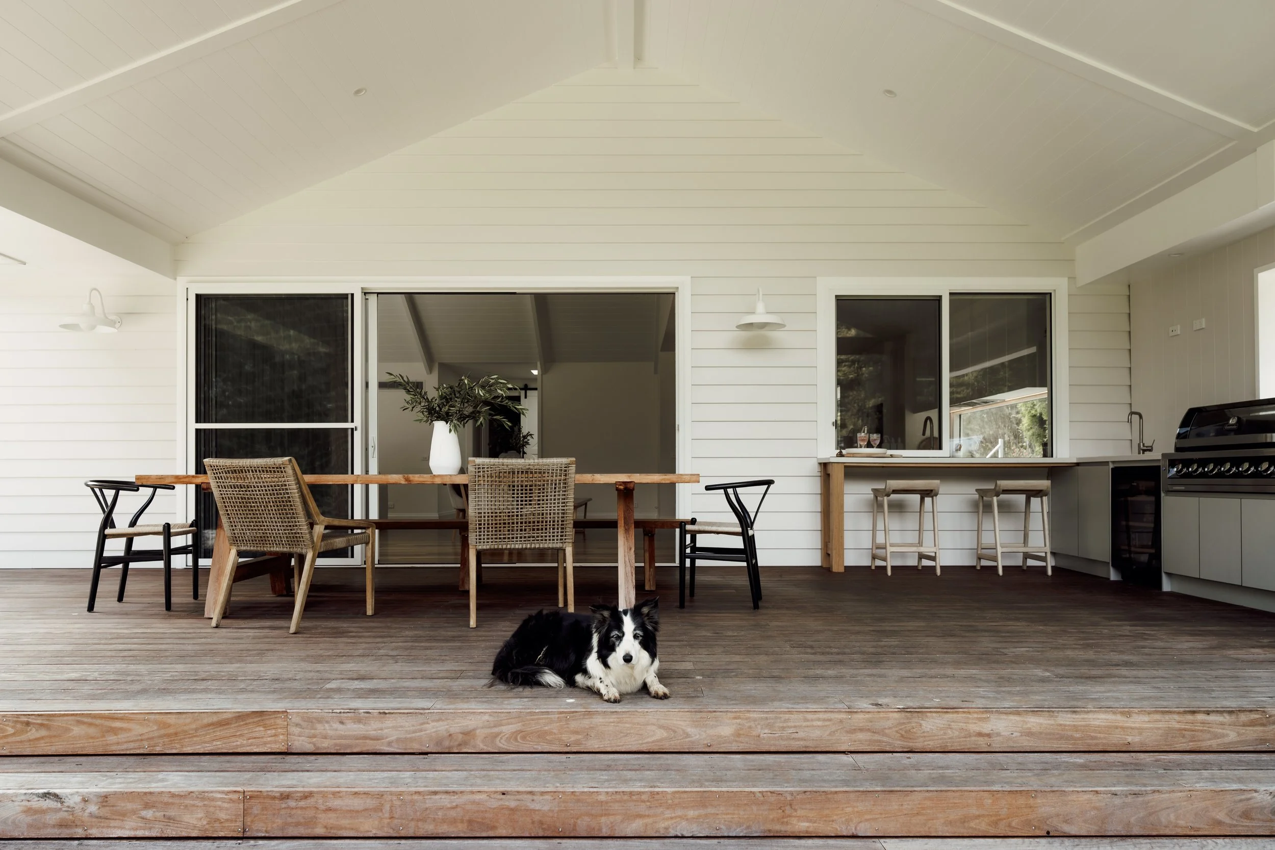 An outdoor patio with a wooden deck, a dining table with chairs, a potted plant, and a dog lying on the floor.