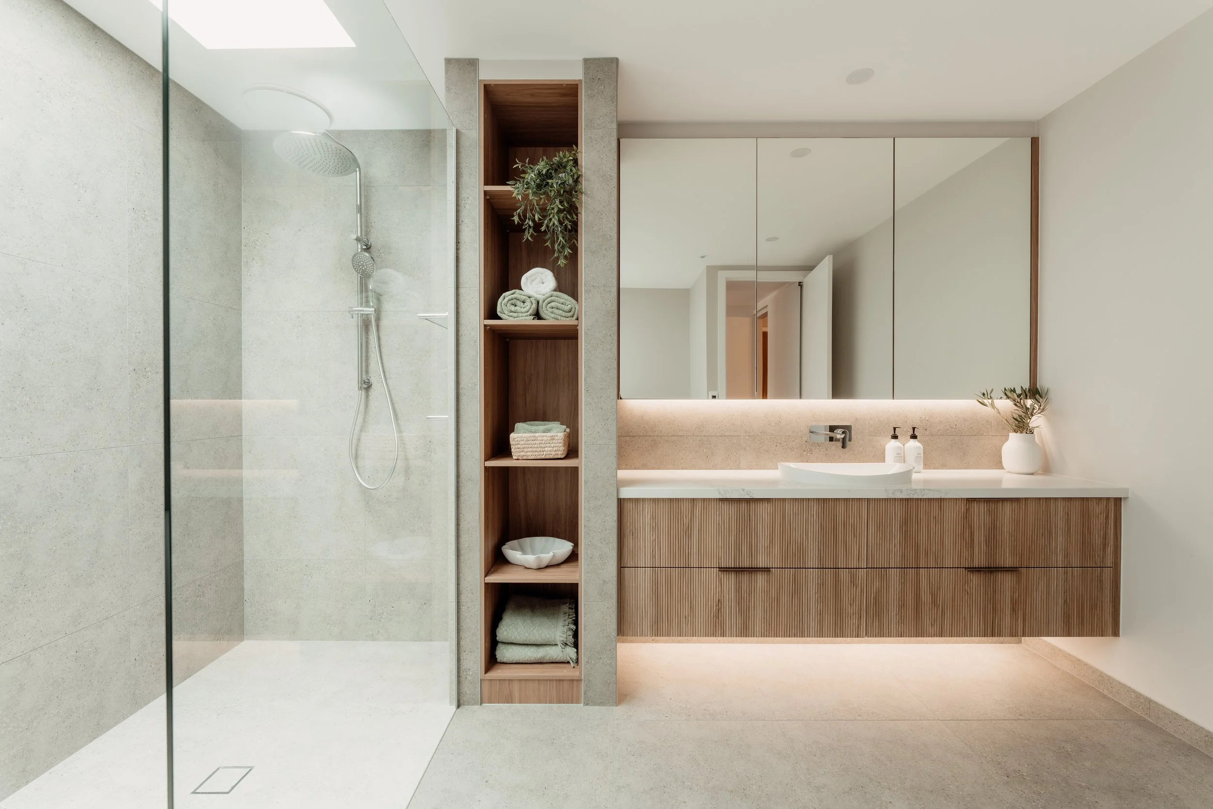 Modern minimalist bathroom with clear glass shower, open shelving with towels and plant, large mirror, light-colored countertop, and wooden cabinet.
