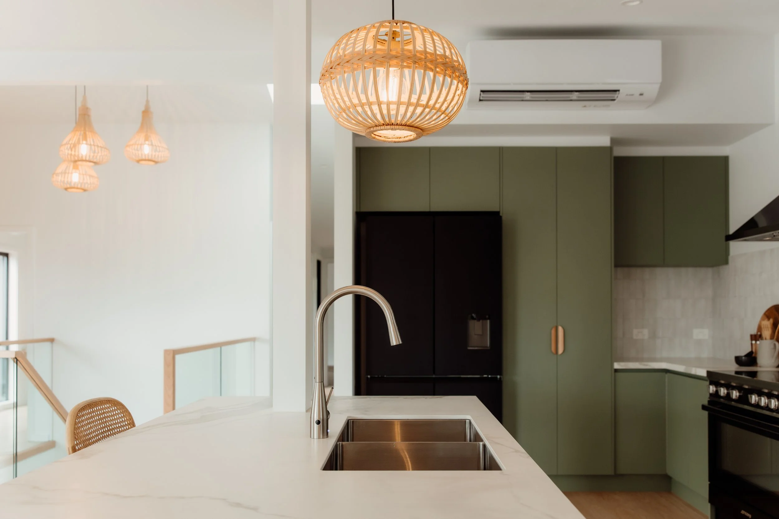 Modern kitchen with white marble countertop, stainless steel sink, black faucet, green and black cabinets, wood accents, and woven pendant lighting.