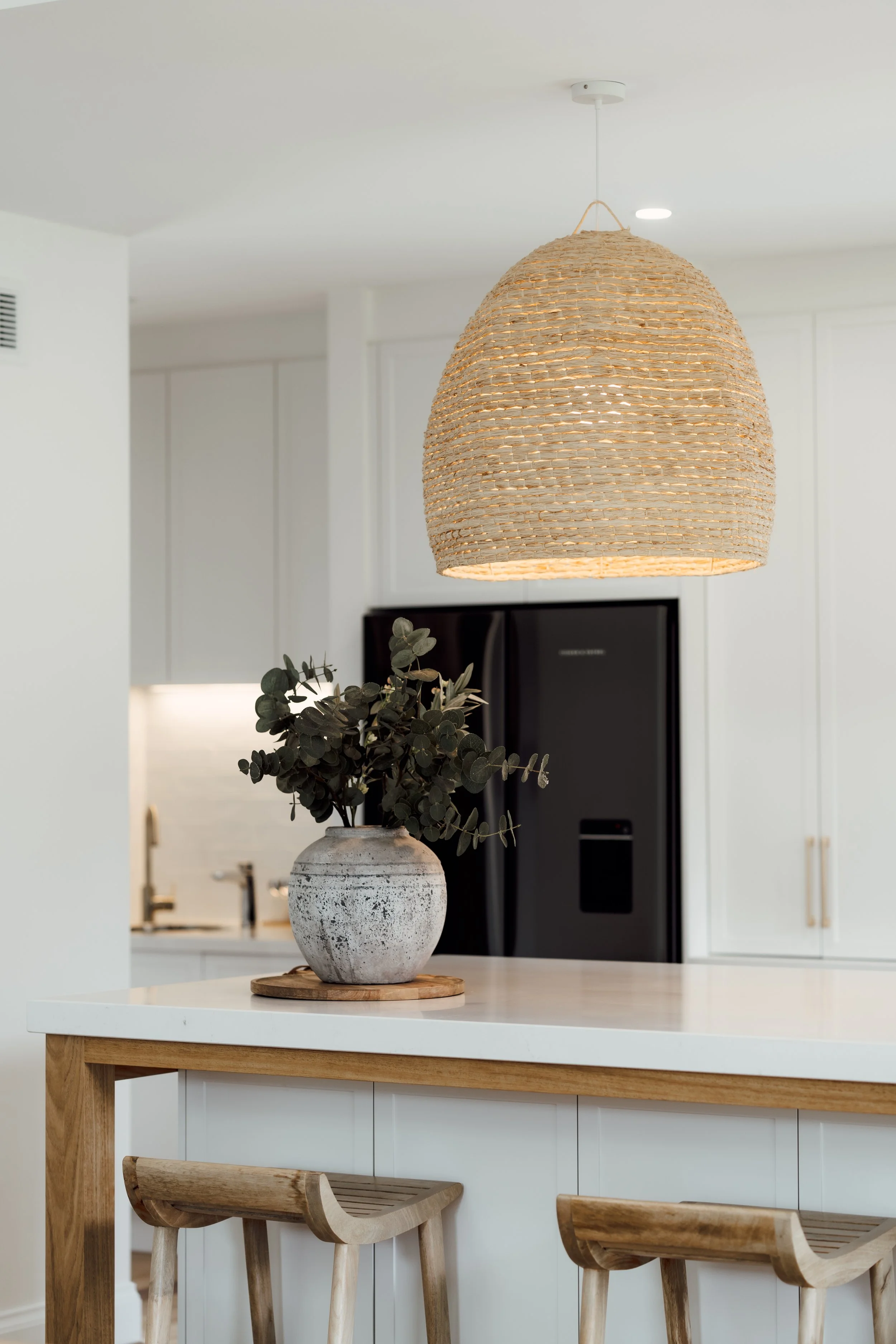 A woven pendant light hanging above a kitchen island with a vase and greenery, in a modern white kitchen with wooden accents.