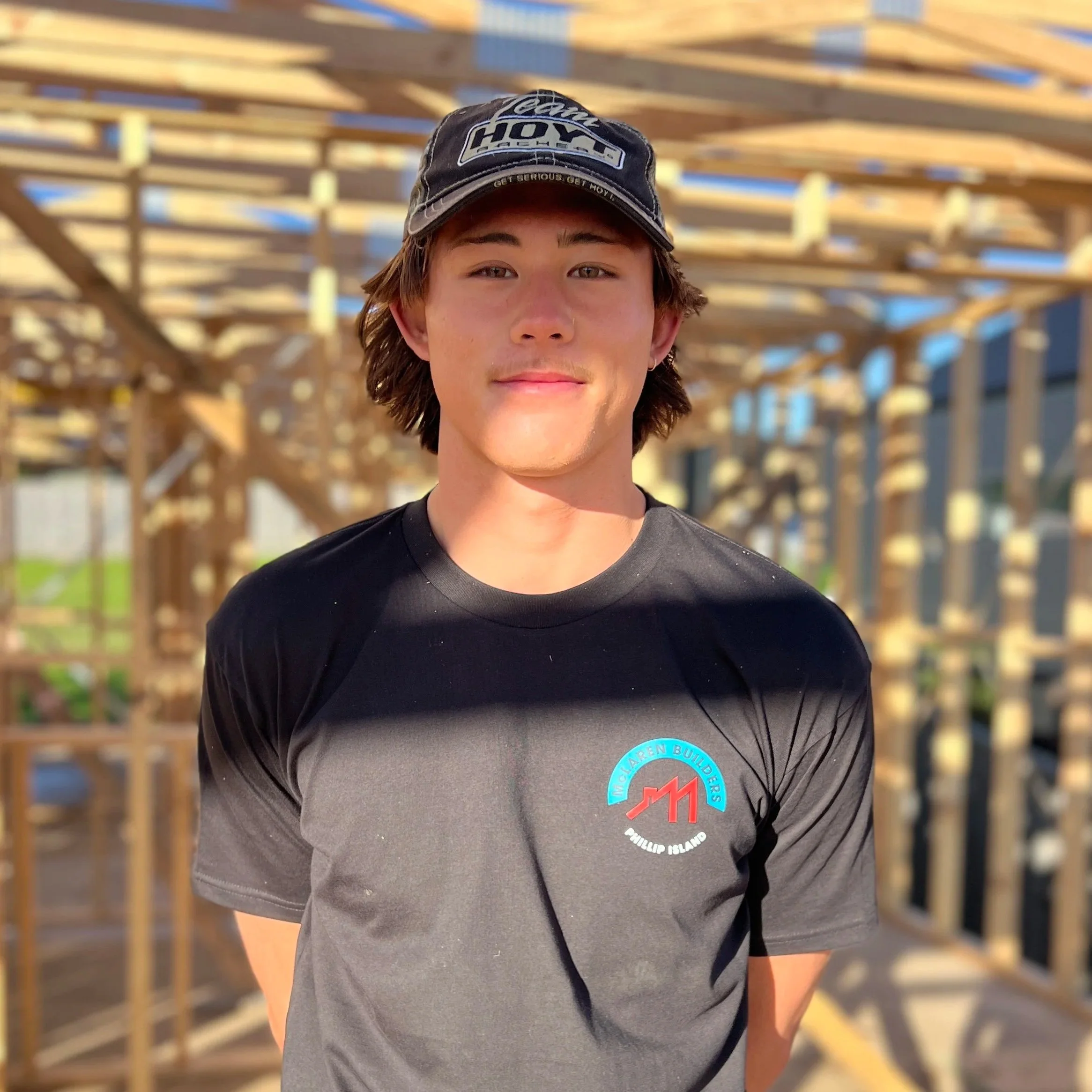 Close-up of a young man wearing a black baseball cap with sponsors logos, standing outdoors in front of a wooden construction framework under blue sky