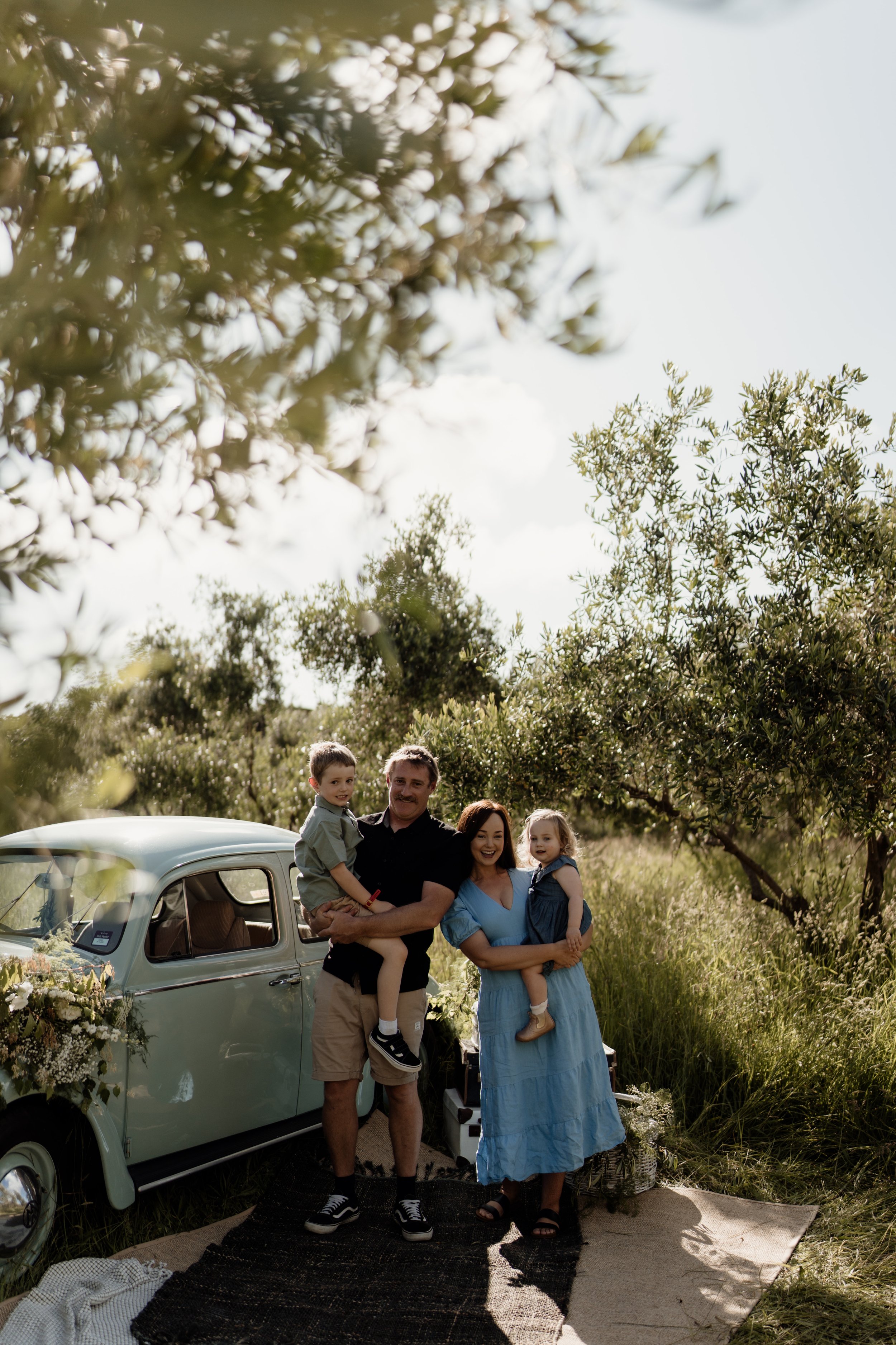 A family of four standing in front of a vintage light blue car decorated with flowers, outdoors, surrounded by trees and grass, with a patterned rug on the ground and baskets filled with flowers nearby, during daytime.