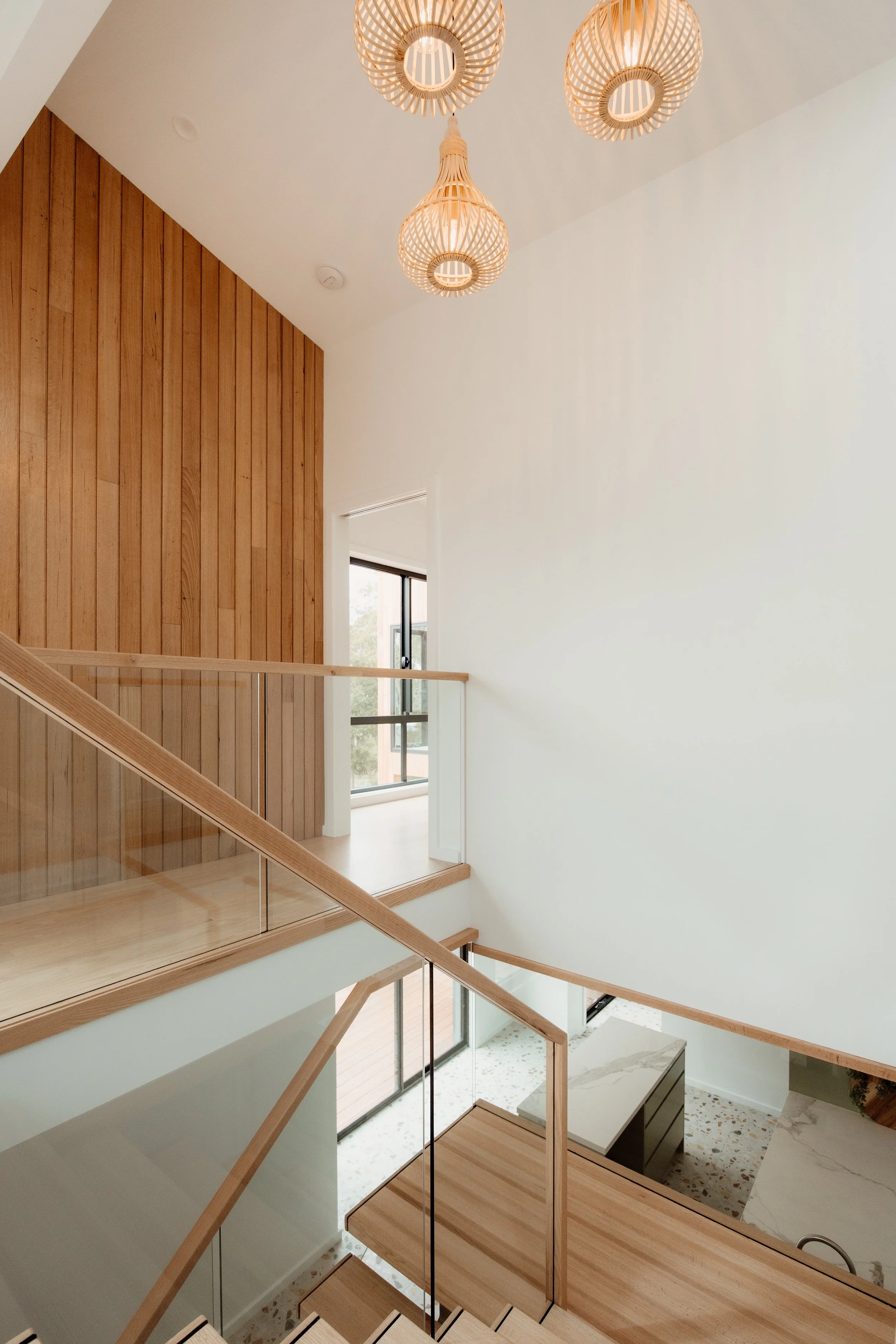 Interior view of a modern home with a wooden staircase, hanging pendant lights, a window, and a mix of white walls and wood paneling.