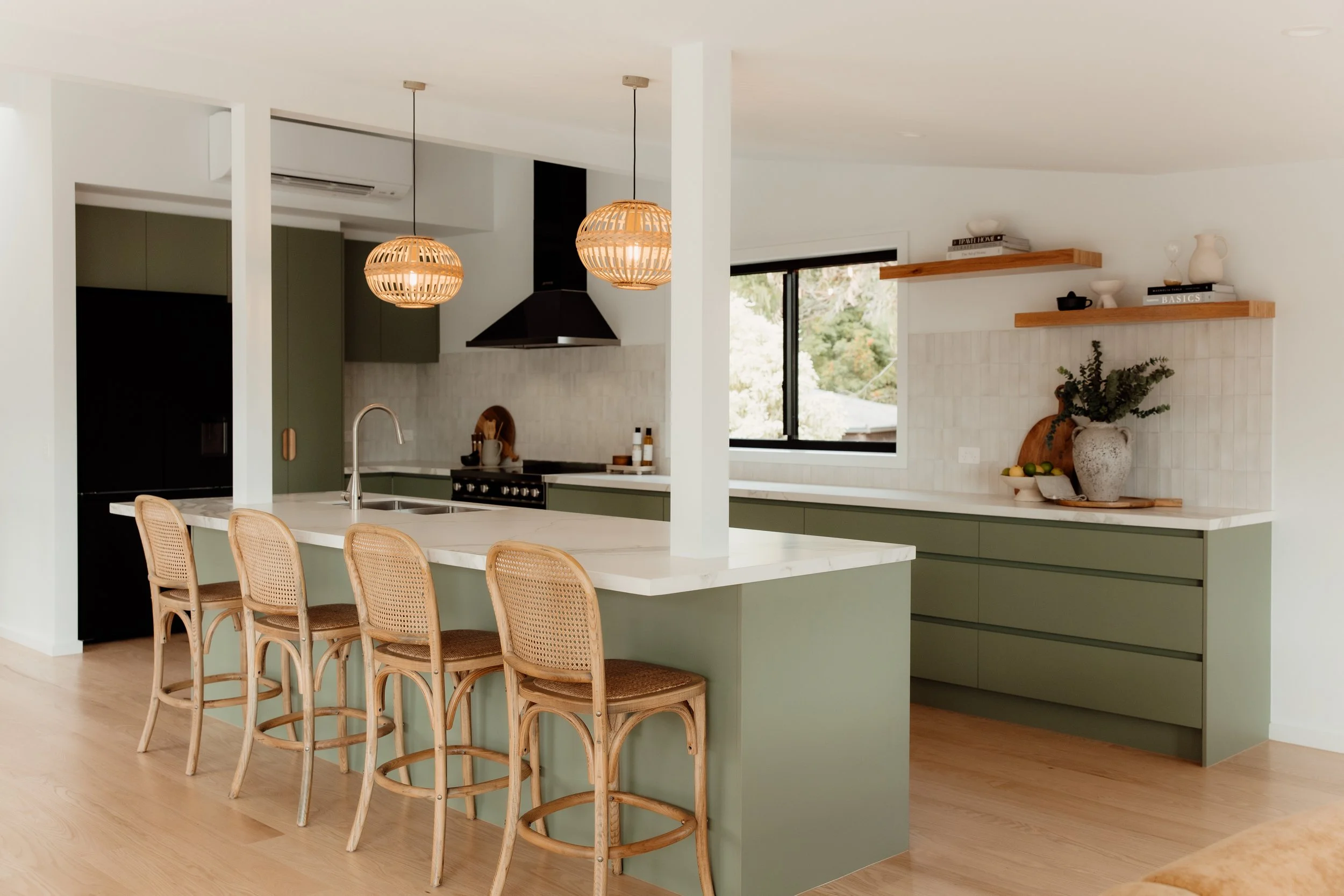 A modern kitchen with a white island counter, wooden bar stools, green cabinets, black range hood, hanging wicker pendant lights, and open wooden shelves with decor and books.
