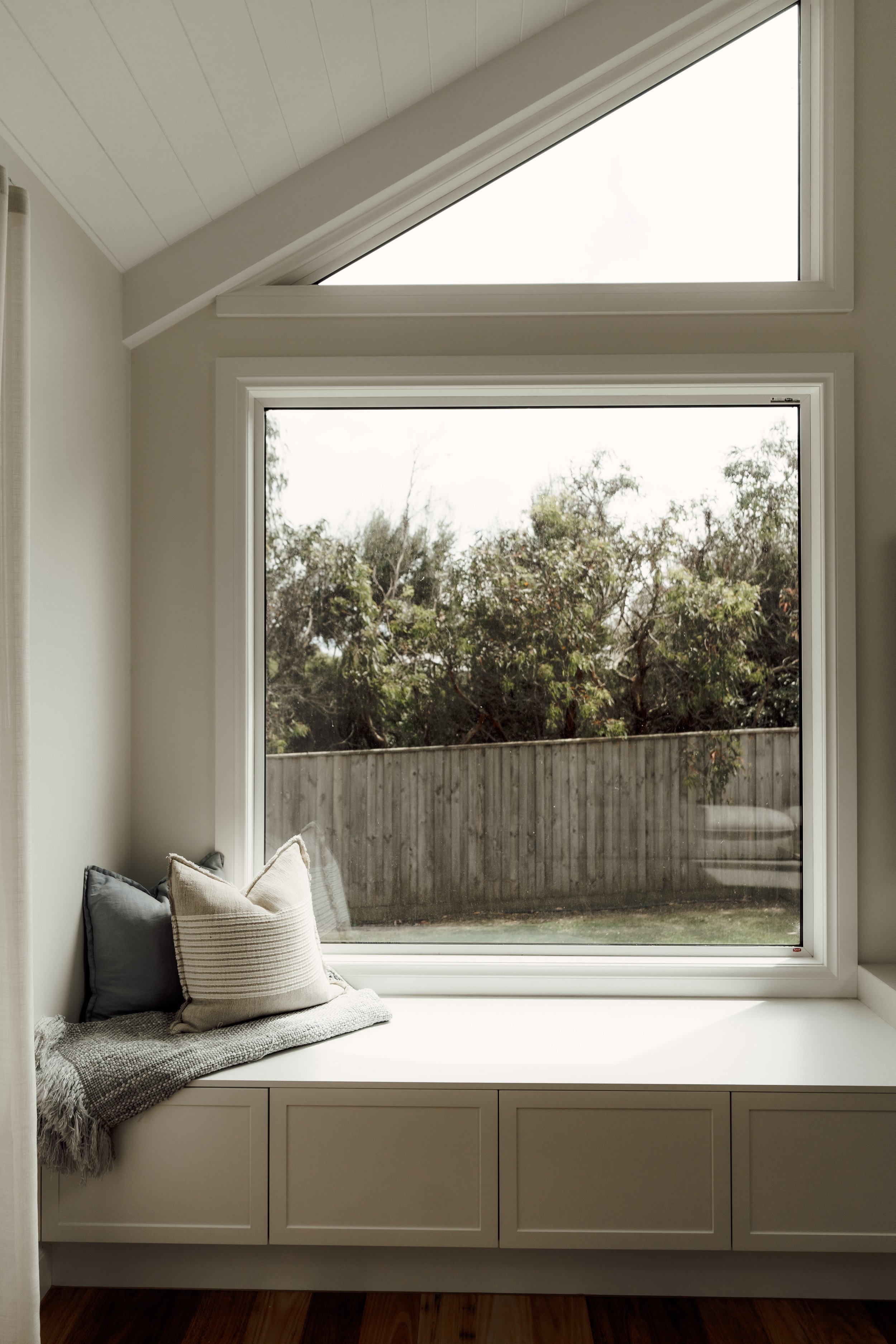 A window seat with pillows and throw blanket in front of a large, angled window showing a backyard with trees and a wooden fence.