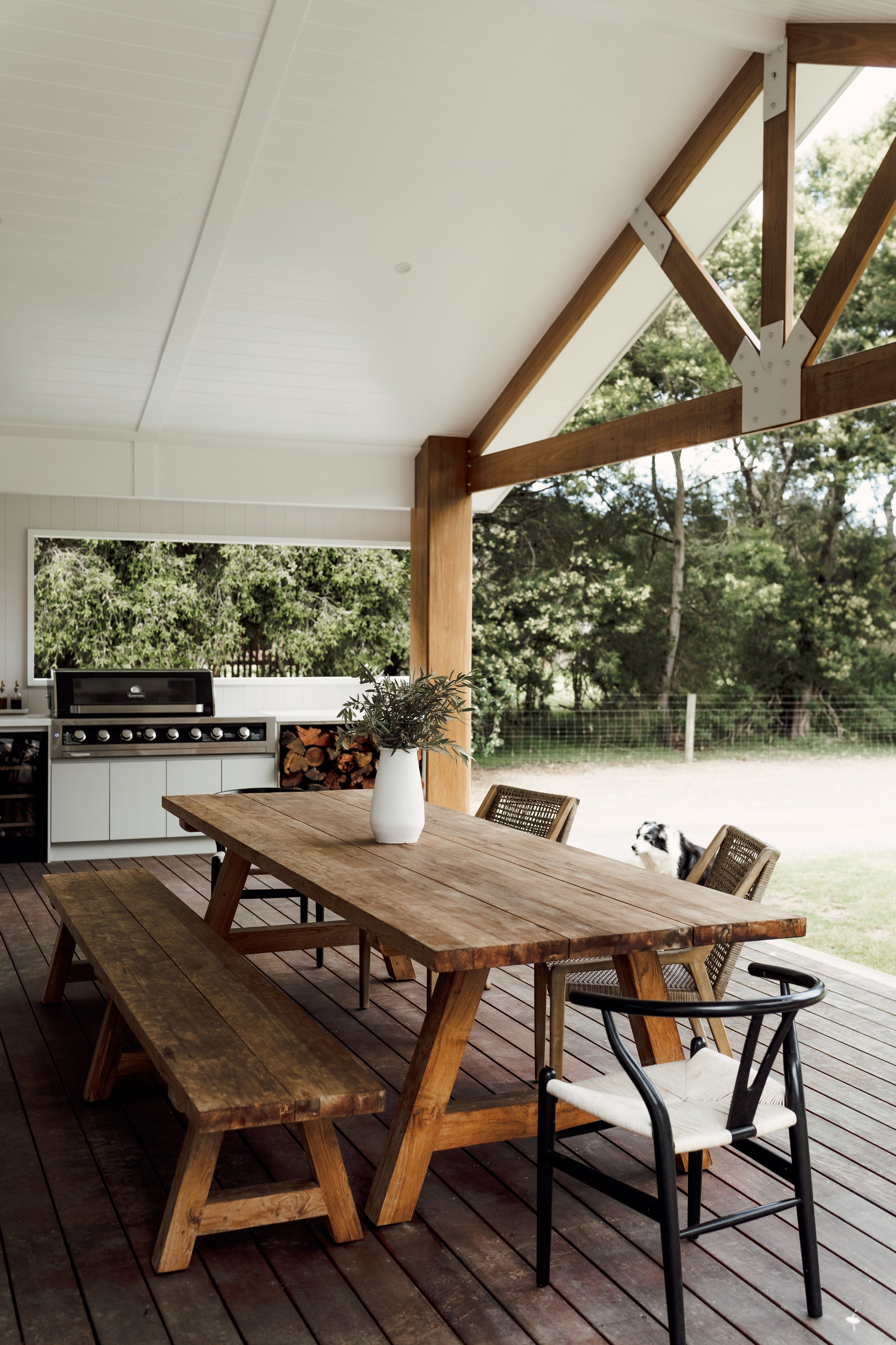 An outdoor patio with a wooden dining table surrounded by a mix of chairs, including a wicker chair and a black metal chair with a white cushion. A white vase with green foliage is on the table. In the background, a black and white dog is near the ya