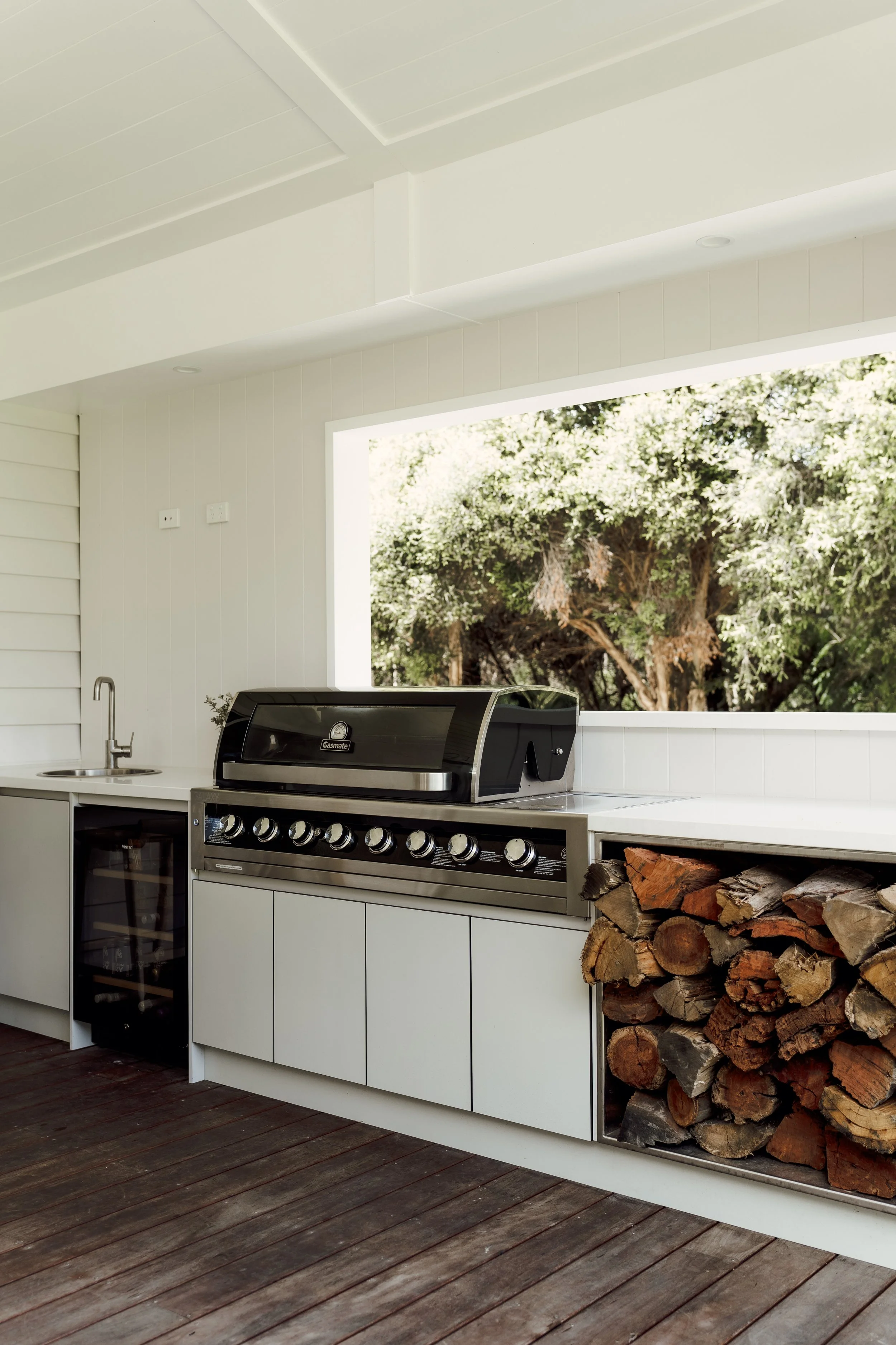 Outdoor kitchen with a stainless steel grill, stacked firewood, white cabinetry, a small sink, and a large window showing trees outside.