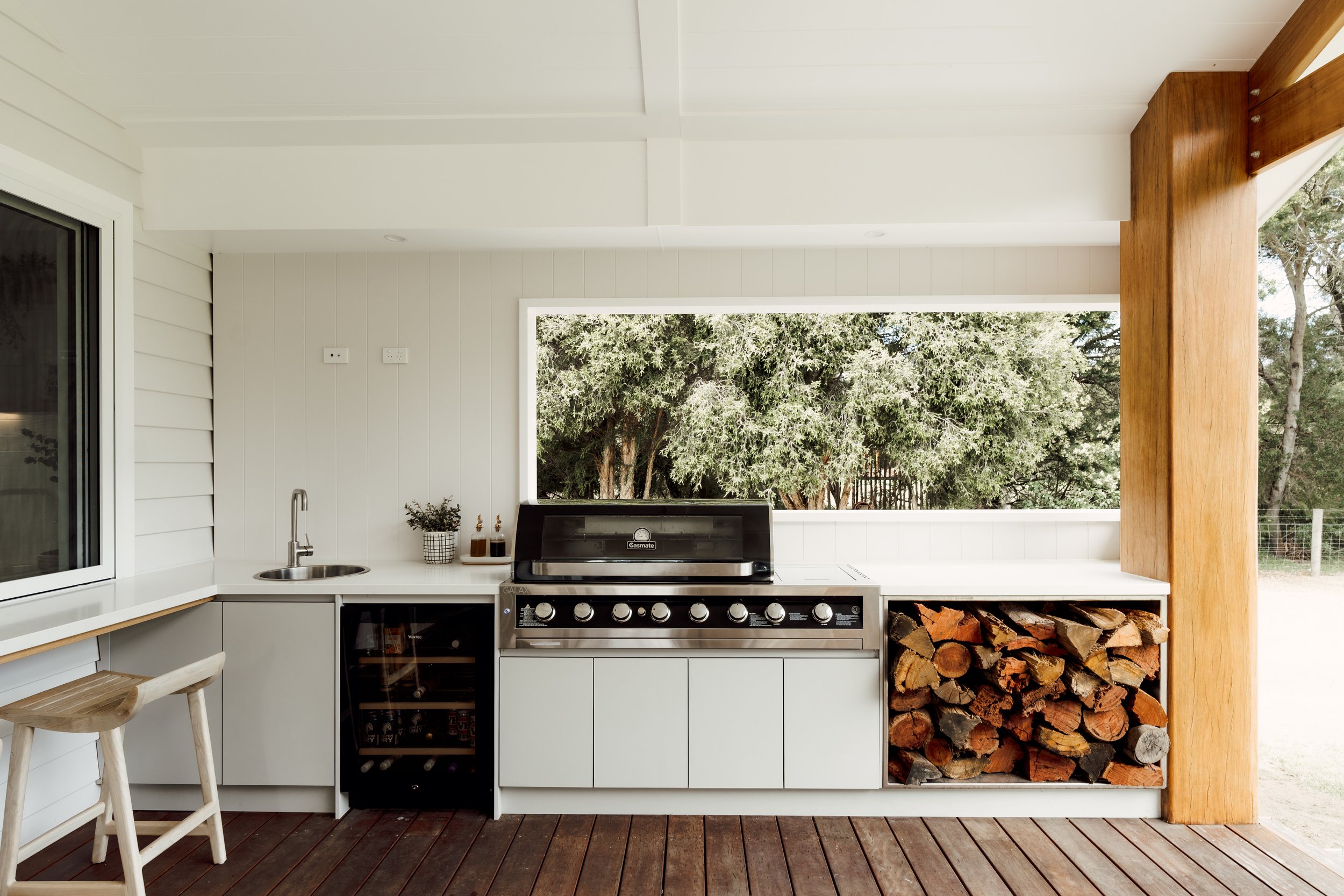 A modern outdoor kitchen with a white countertop, gas grill, wood storage, and large window overlooking trees.