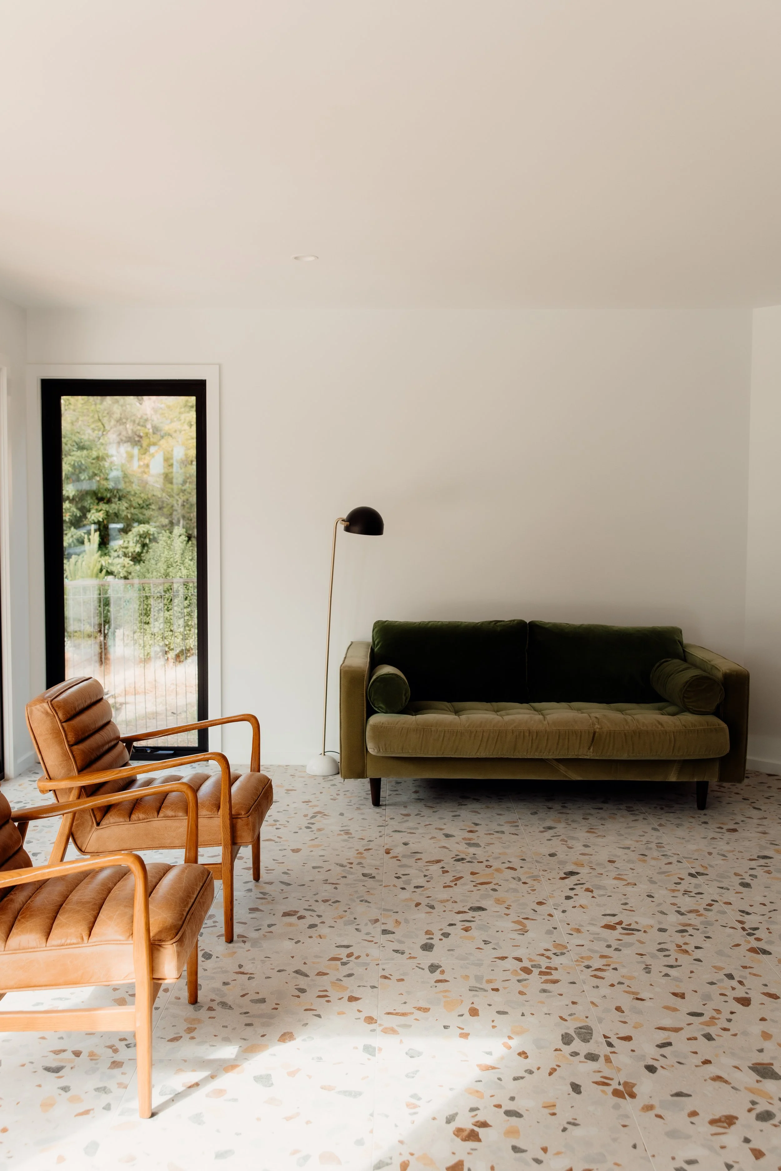 Living room with terrazzo flooring, a green velvet sofa, a black lamp, a wooden armchair with brown leather cushions, and a large window showing greenery outside.