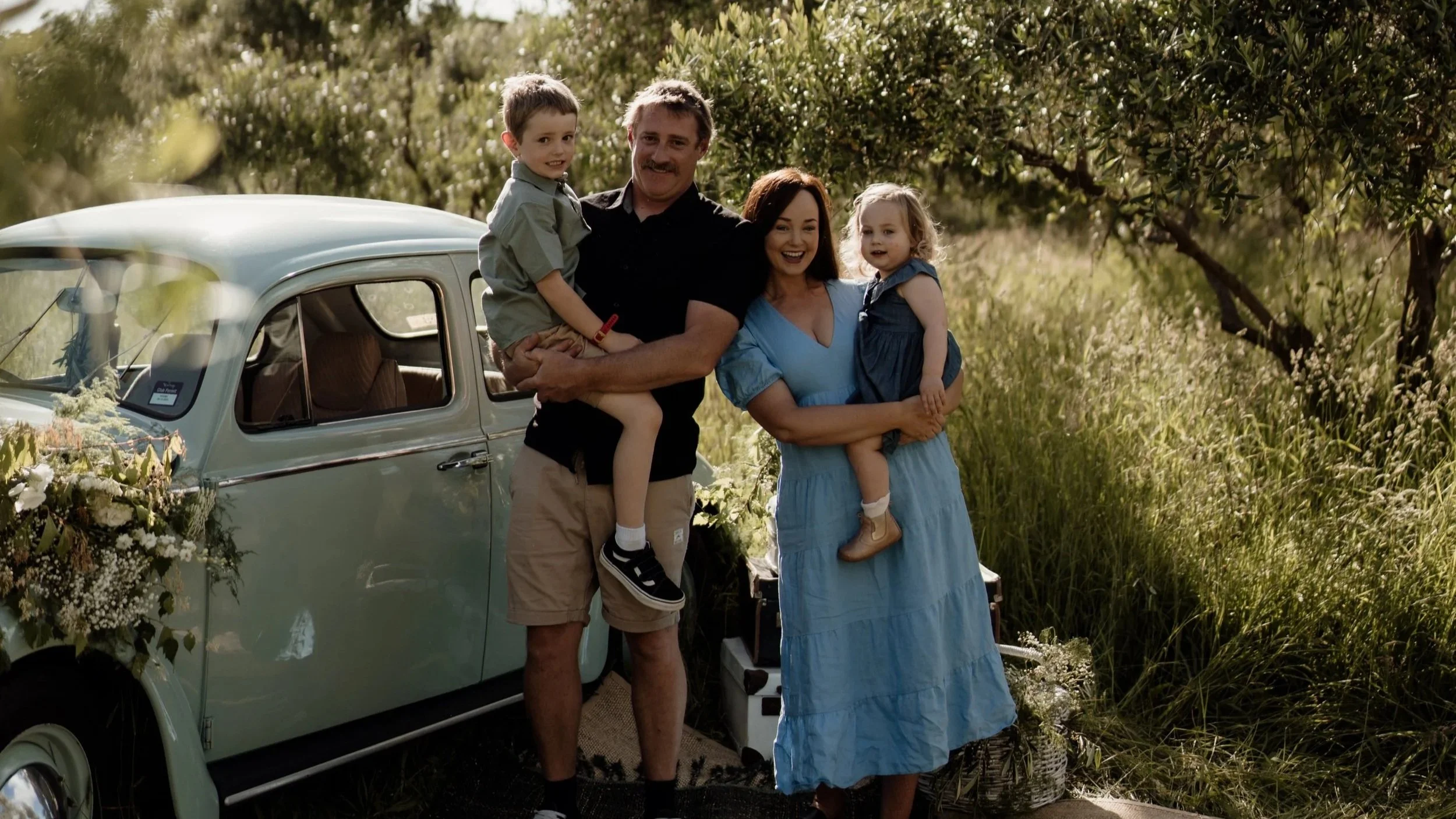 A family of four, including a man, a woman, a boy, and a girl, standing outdoors near a vintage light blue car decorated with flowers. They are smiling and holding each other, with trees and greenery in the background during the daytime.
