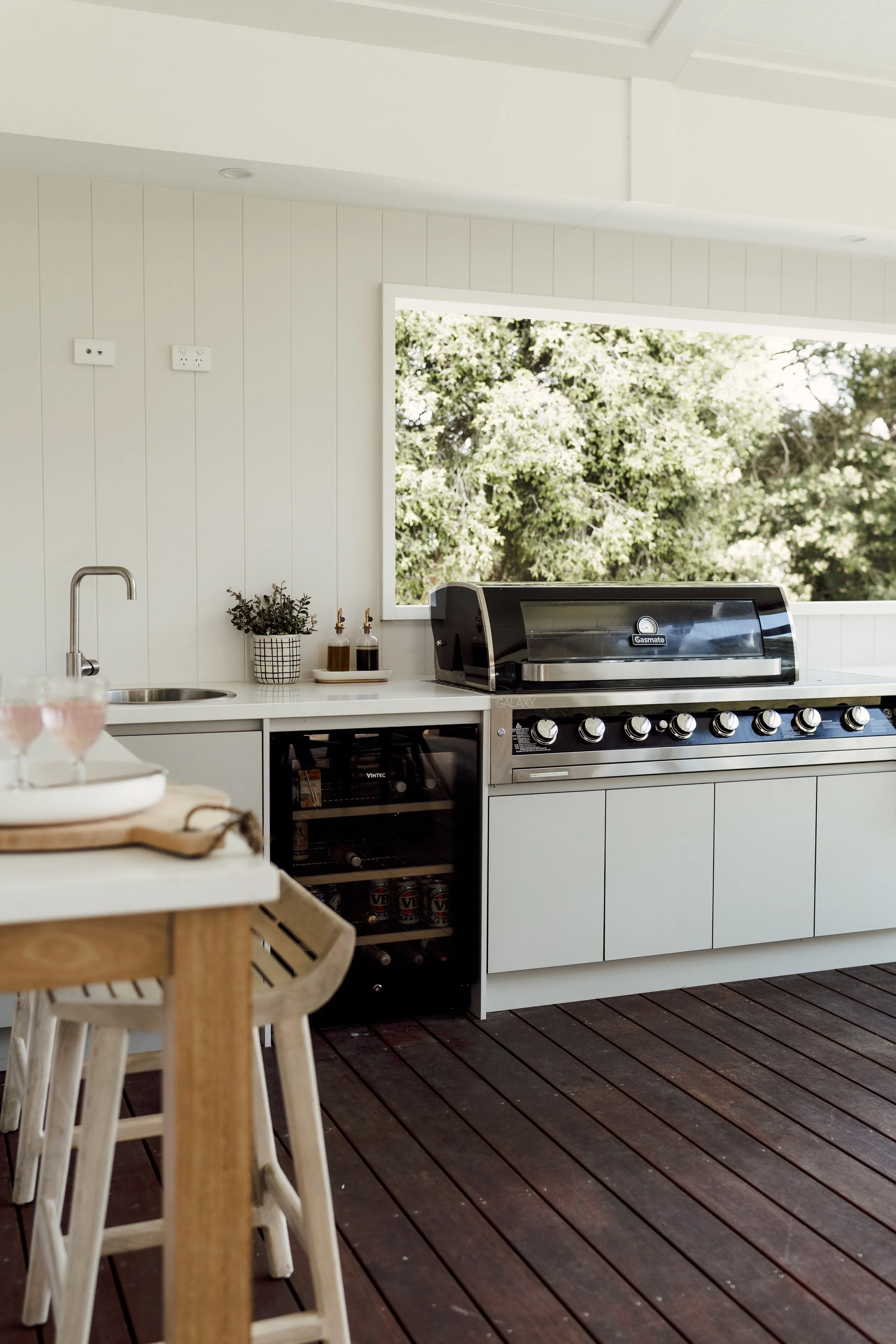 An outdoor kitchen area with a white countertop, a grill, and a window showing green trees outside. There is a potted plant and soap dispensers on the counter, and a wooden table with chairs is partially visible in the foreground.