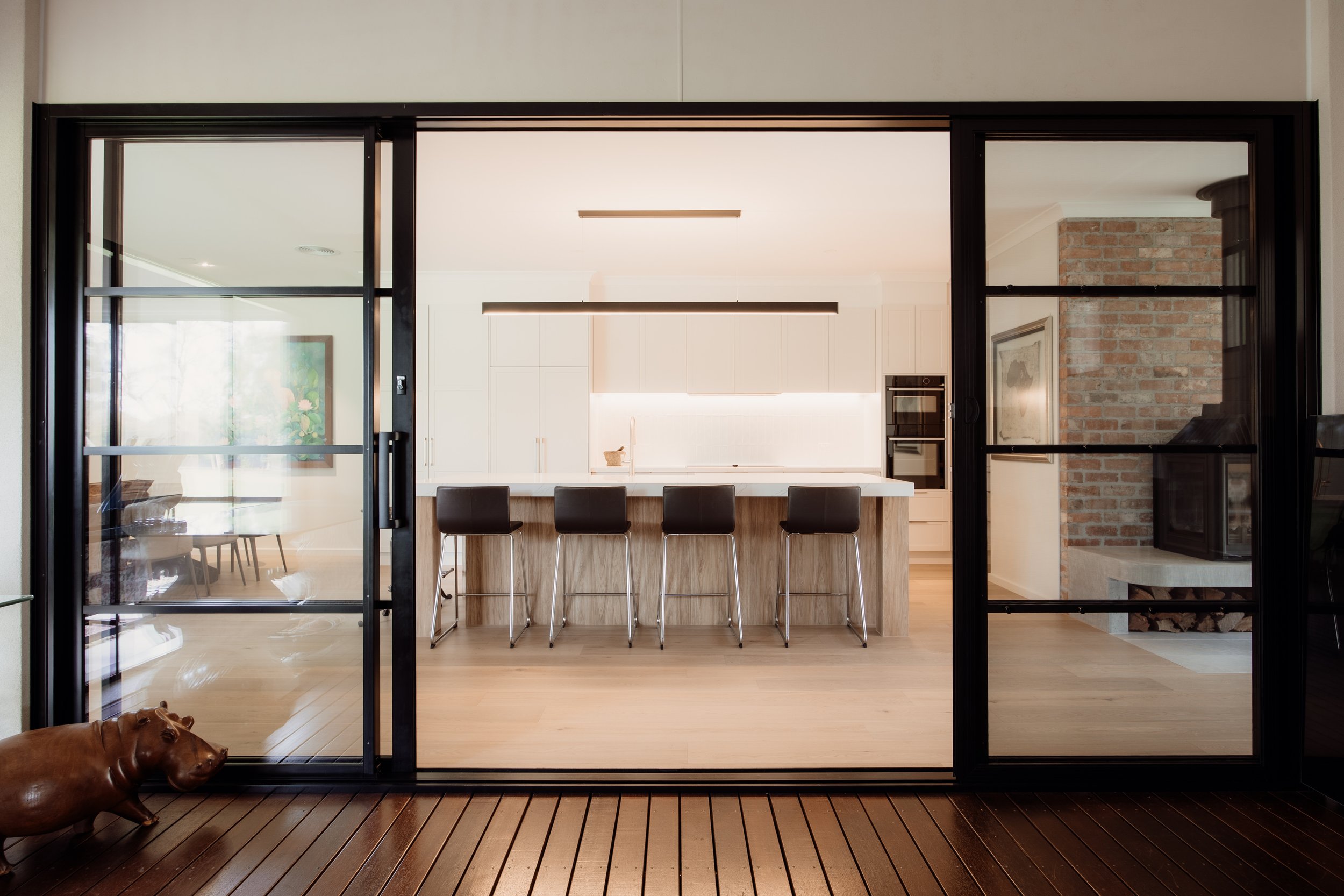 View through glass sliding doors into a modern kitchen with a wooden island and five black bar stools, brick wall on the right, white cabinets, and a built-in oven; a brown dog is lying on wooden deck outside.