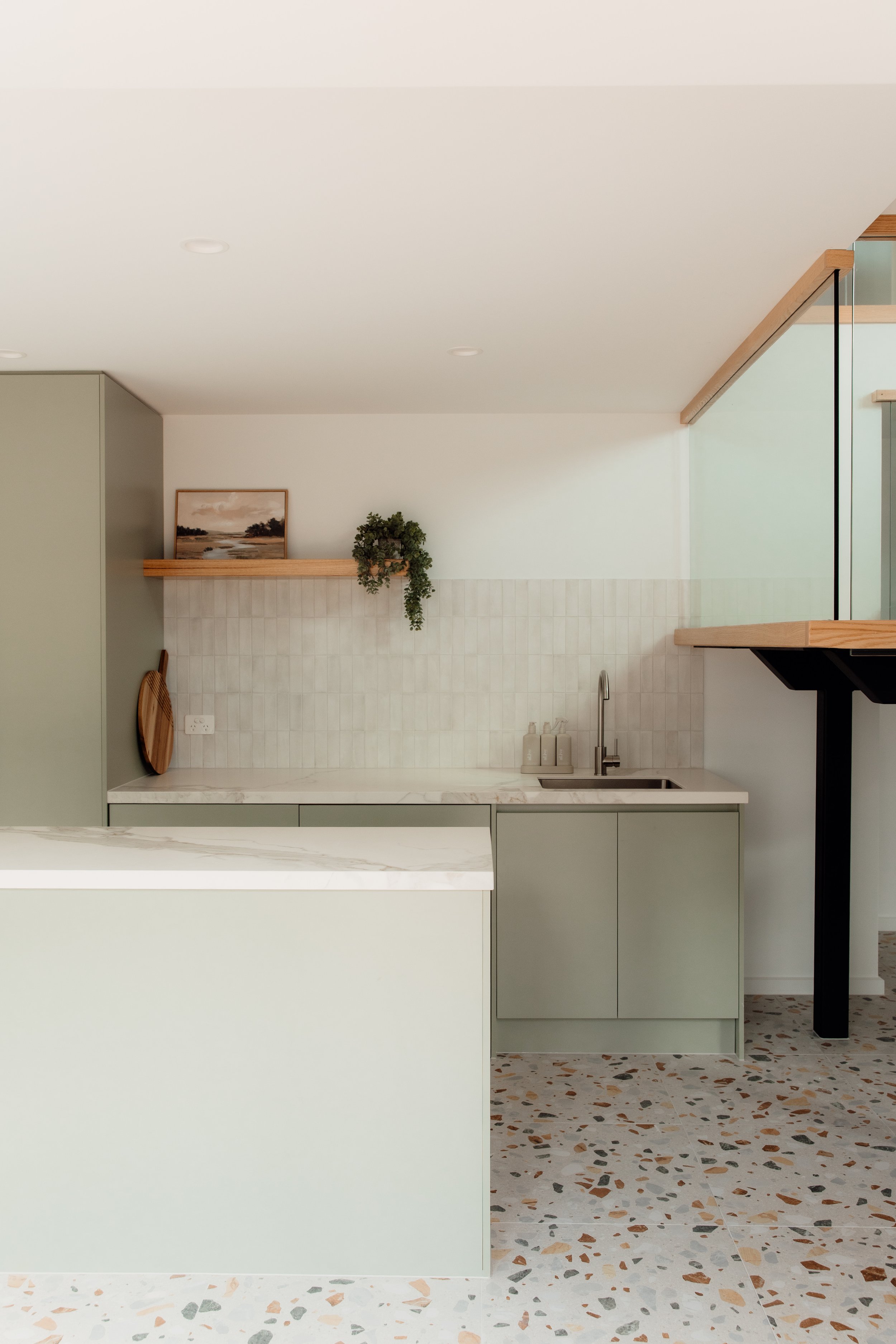 Modern kitchen with light green cabinets, a marble countertop, a sink with a faucet, and terrazzo tile flooring. A wooden shelf holds a landscape painting and a potted plant.