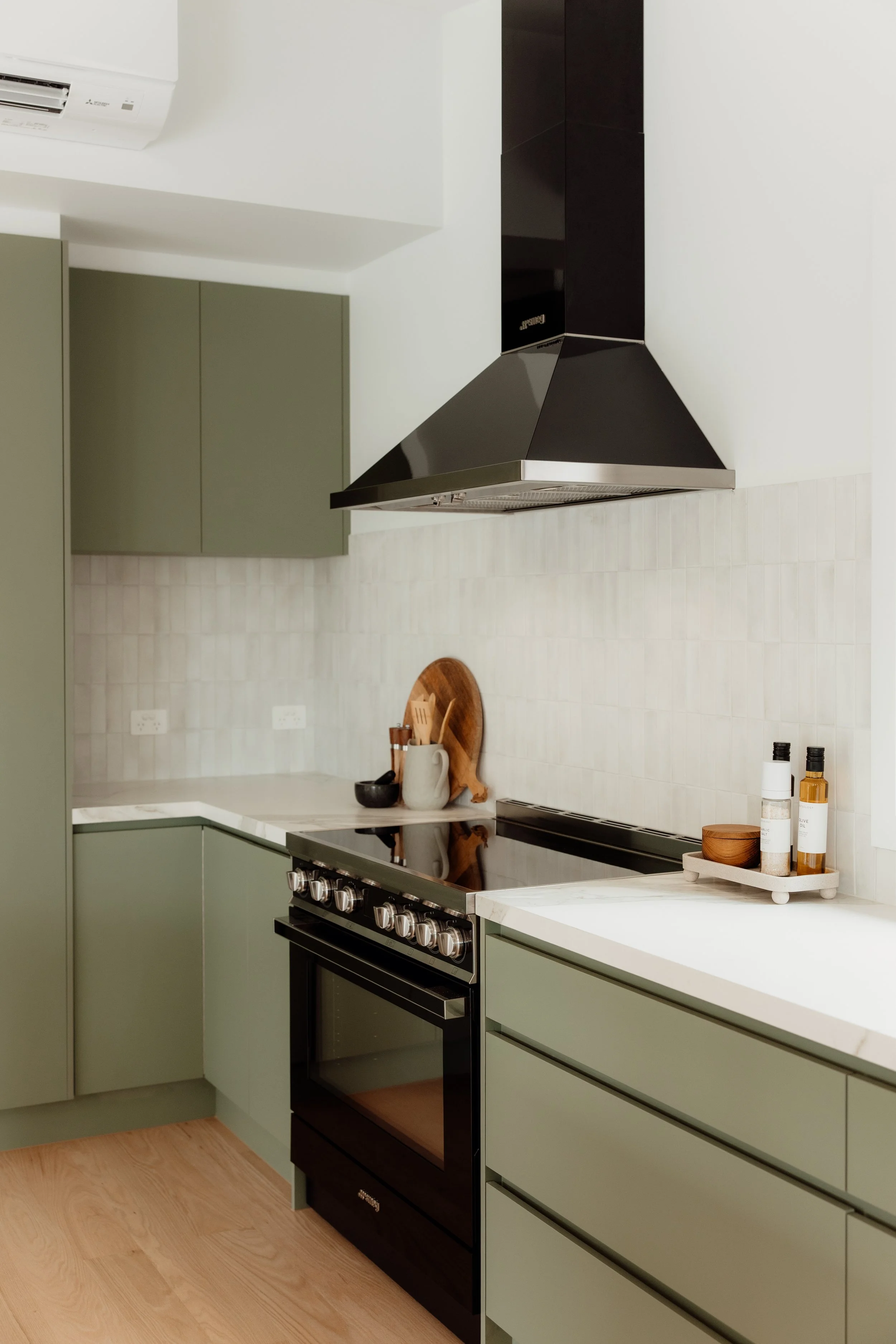 Modern kitchen with black oven, green cabinetry, white marble countertop, and a black range hood. Small cutting board and bottles of oil or vinegar on a white tray.