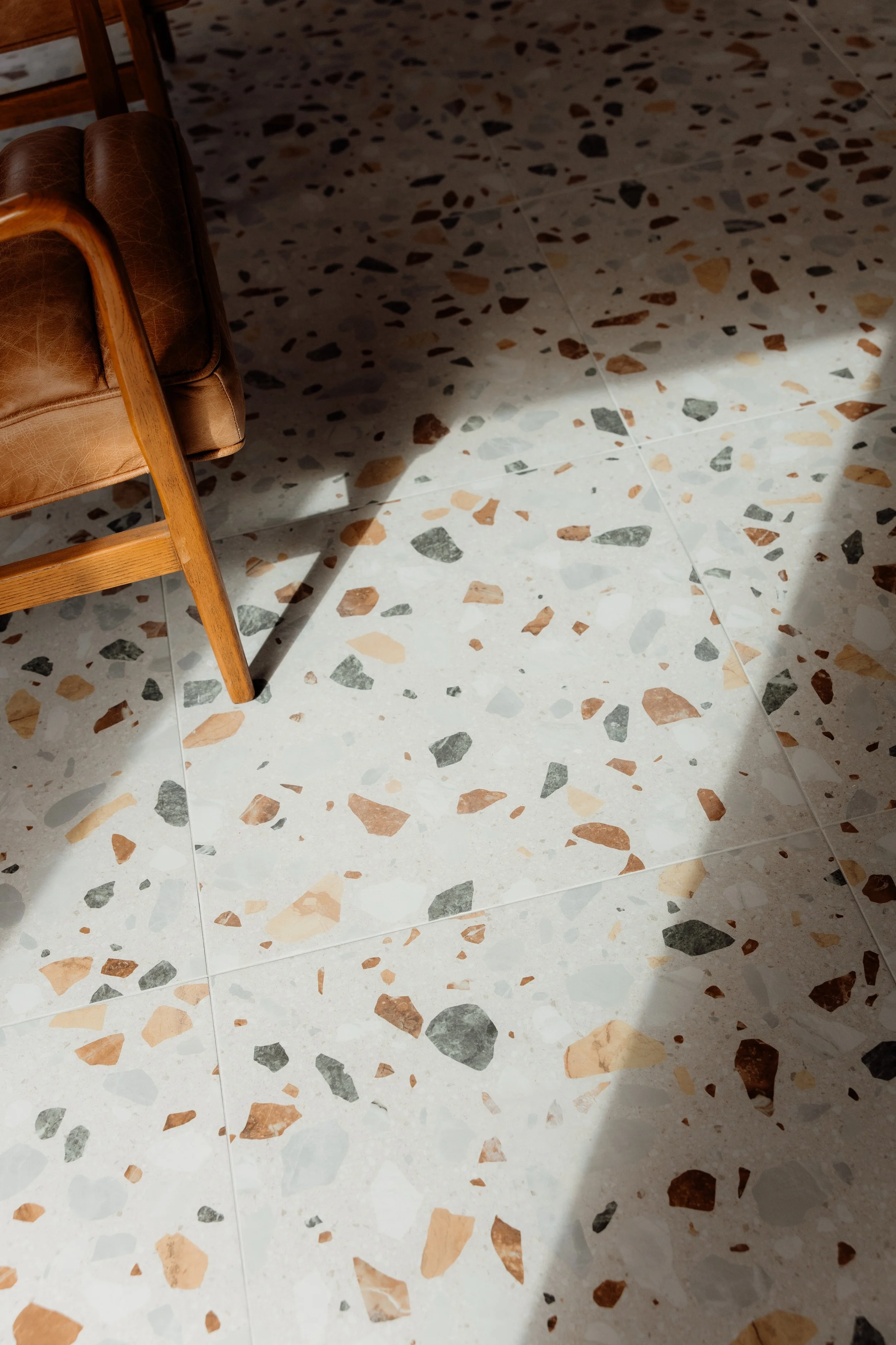 Part of a brown leather armchair with wooden legs positioned on a terrazzo floor with multicolored stone chips in a pattern of white, beige, black, and brown.
