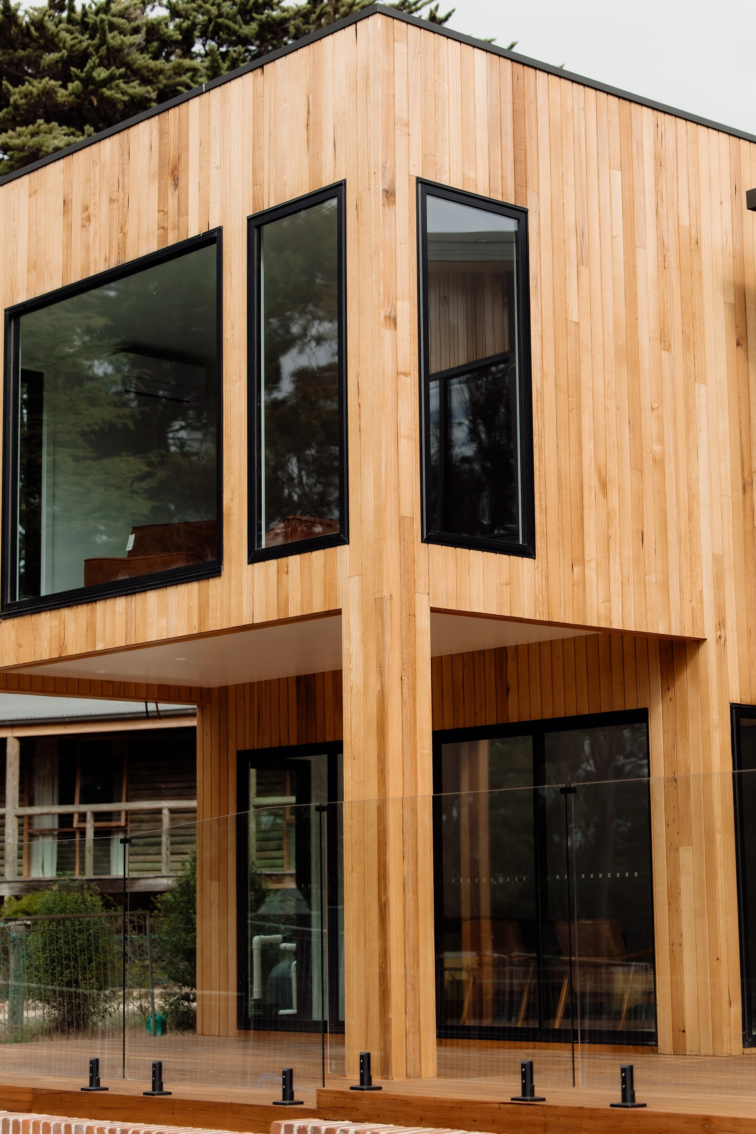 Modern house with wooden exterior and large black-framed windows, balcony with glass railing, surrounded by trees.