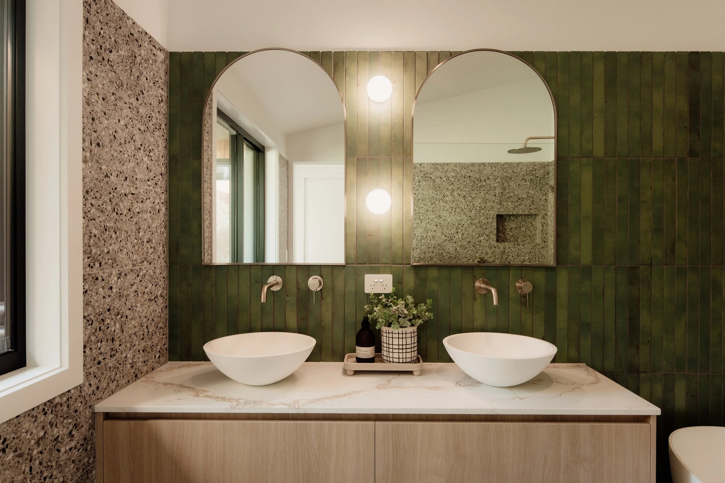 A bathroom vanity with two white vessel sinks, a marble countertop, and green tiled walls with mirrors, round lights, and decorative items.
