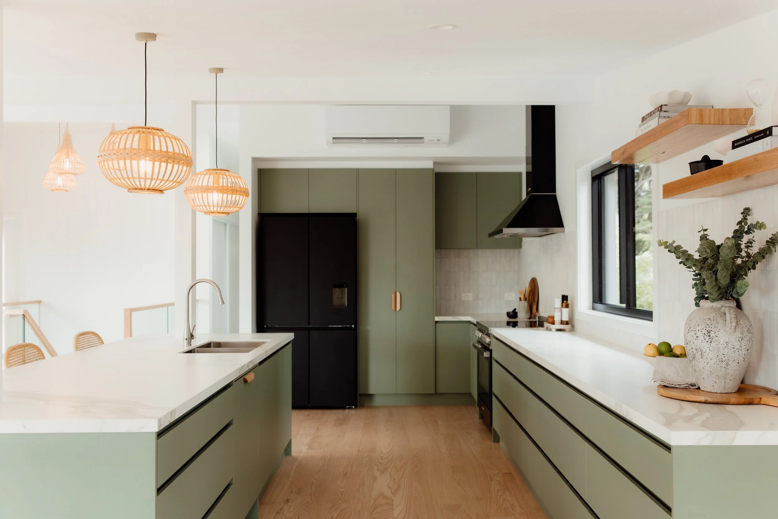 Modern kitchen with green cabinets, white marble countertops, hanging rattan pendant lights, black refrigerator, black range hood, large window with view outside, open shelving with books and ceramics, decorative vase with greenery.