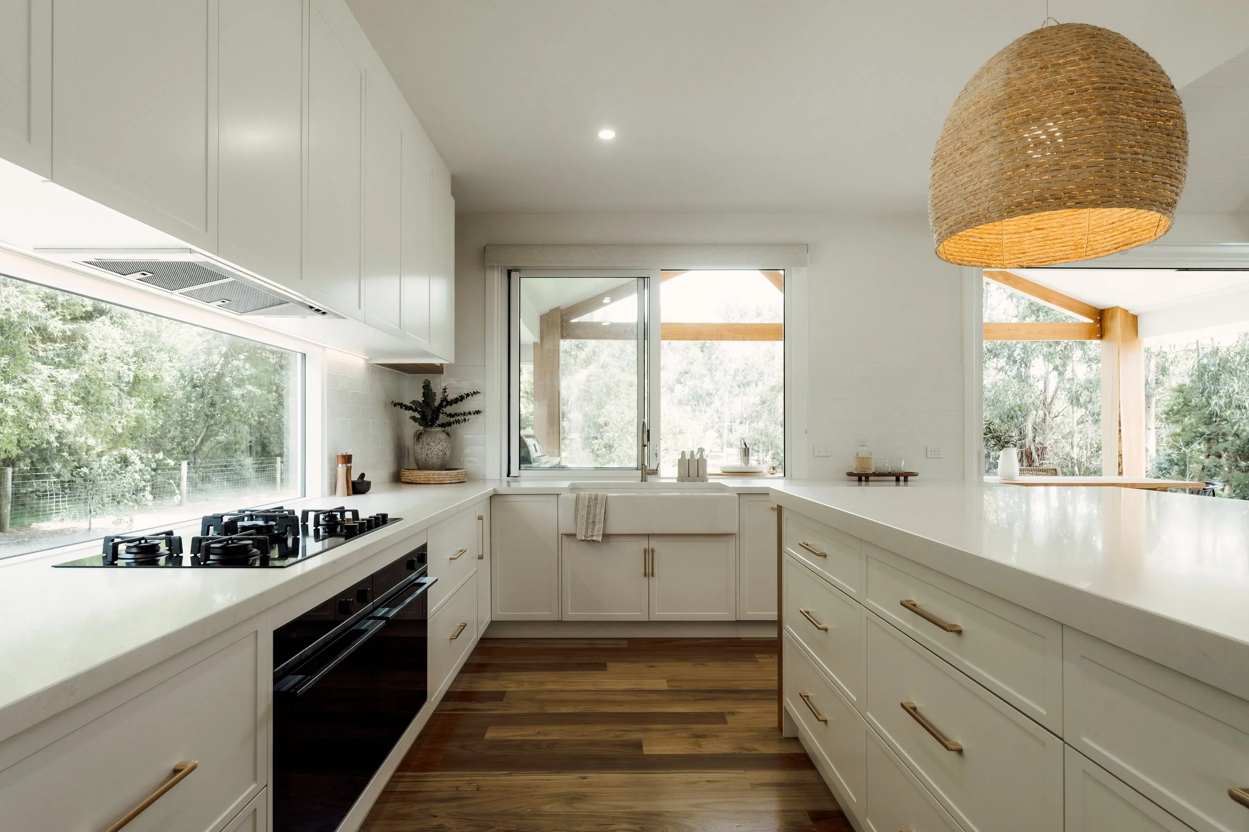 A modern kitchen with white cabinets, a white countertop, large windows showing greenery outside, and a woven pendant light hanging from the ceiling.