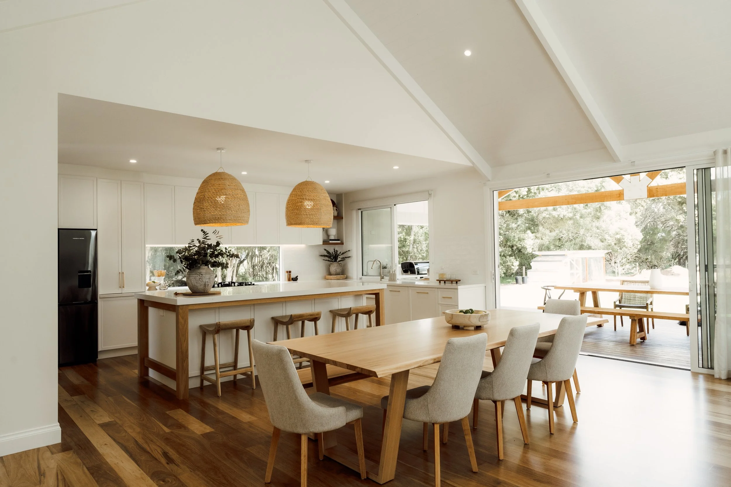 Modern kitchen and dining area with large windows, white cabinets, wooden dining table, and woven pendant lights.