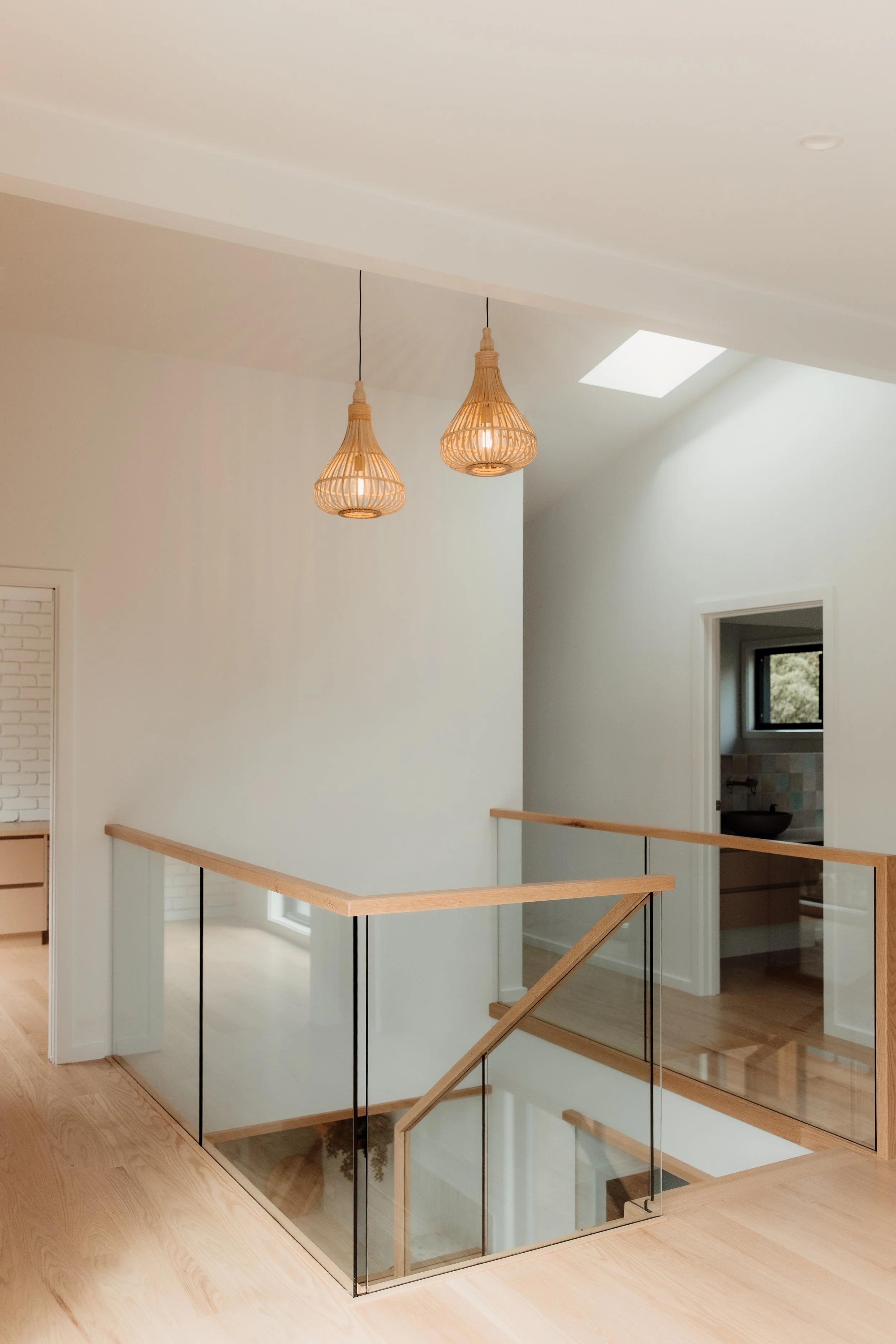 Interior view of a modern home staircase area with wooden flooring, glass railing, and hanging wicker pendant lights, featuring skylights and a door leading to a bathroom.