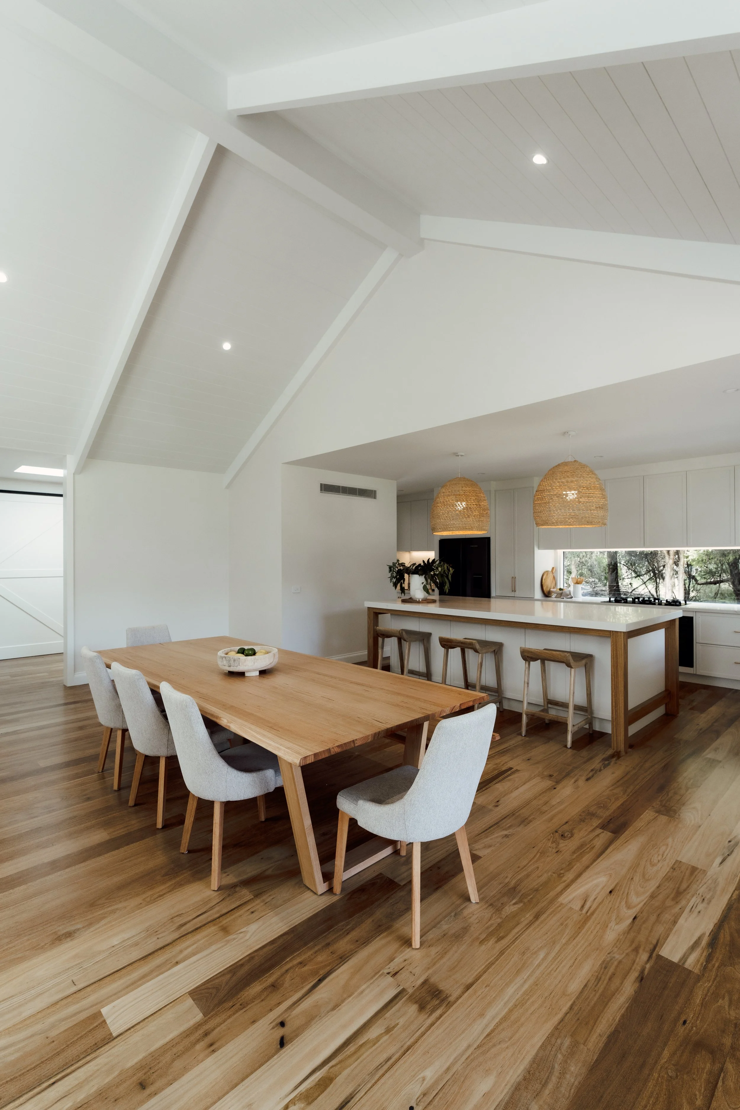 A modern kitchen and dining area with white walls and ceiling, wooden flooring, a wooden dining table with six upholstered chairs, a kitchen island with three stools, hanging wicker pendant lights, and a window overlooking greenery.