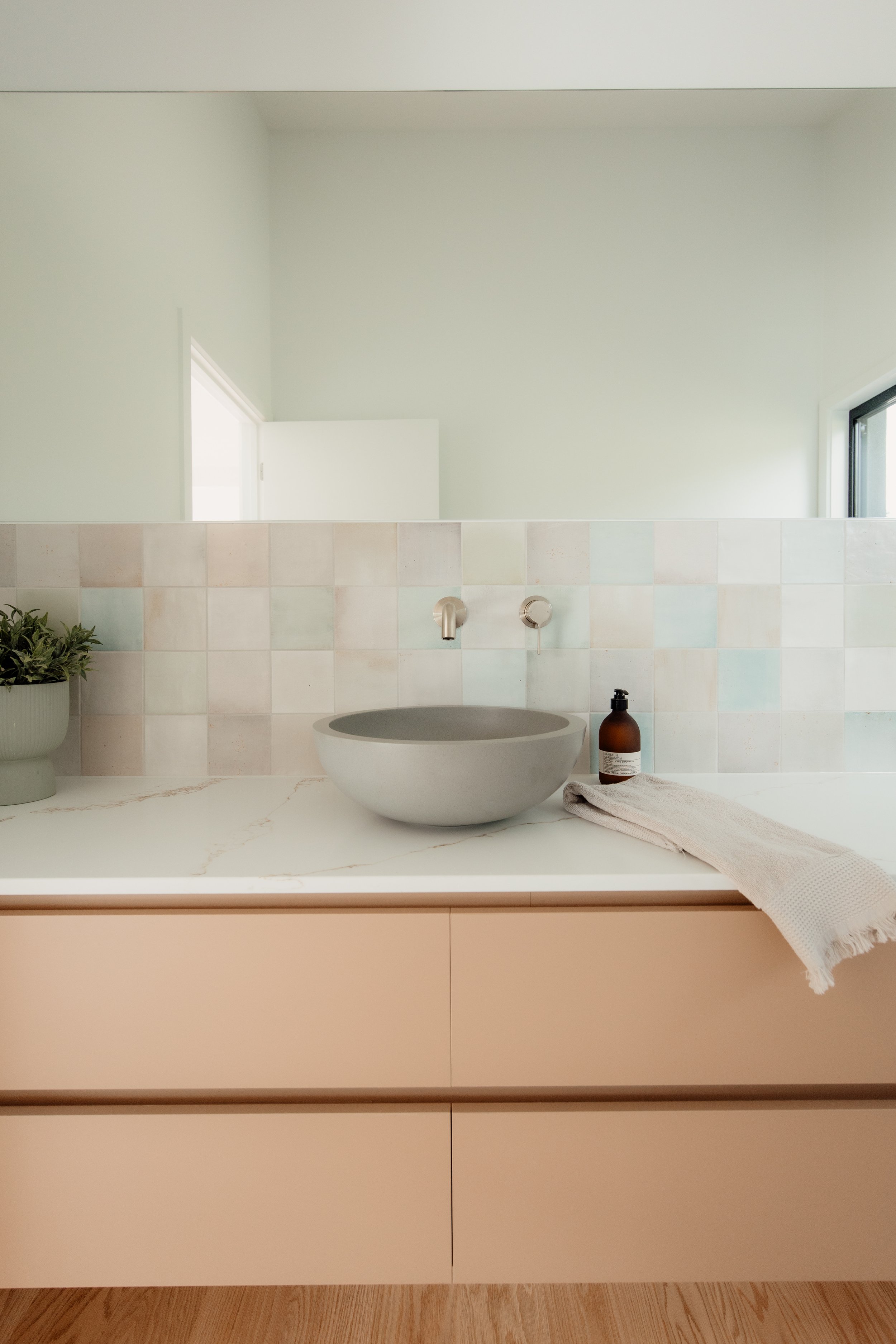 A modern bathroom counter with a large round vessel sink, a soap dispenser, and a beige towel. The background features two faucets, a tiled backsplash, a minimal mirror, and a window.