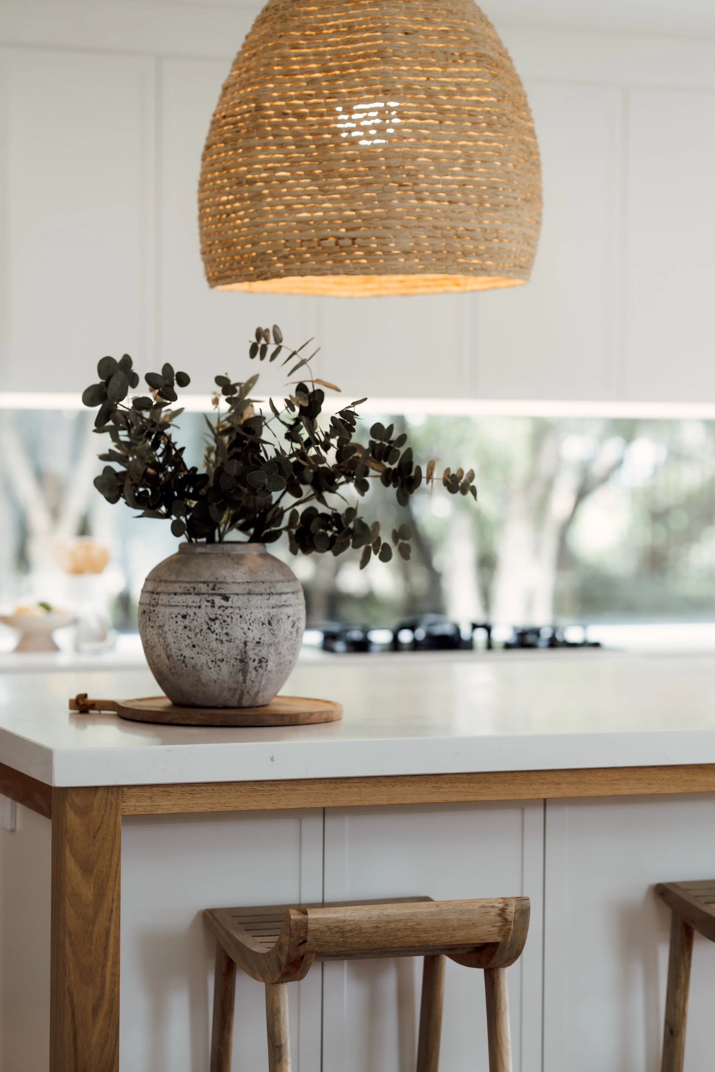 A kitchen countertop with a round ceramic vase holding eucalyptus branches, a wooden tray underneath, and a wooden barstool underneath the counter. A woven pendant light hangs above, and a window with blurred trees outside is in the background.
