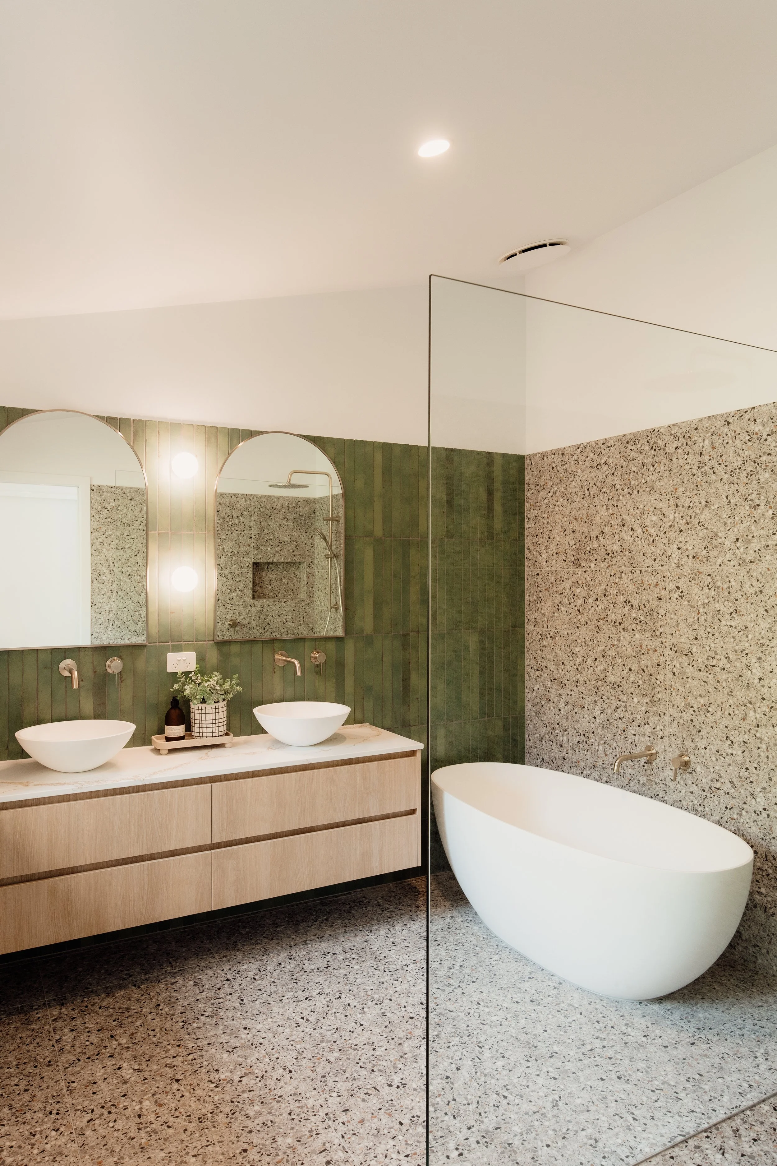 Modern bathroom with a large white freestanding bathtub, green tiled wall, and wooden vanity with two vessel sinks, round mirrors, and potted plant; ceiling light and speckled tile flooring.