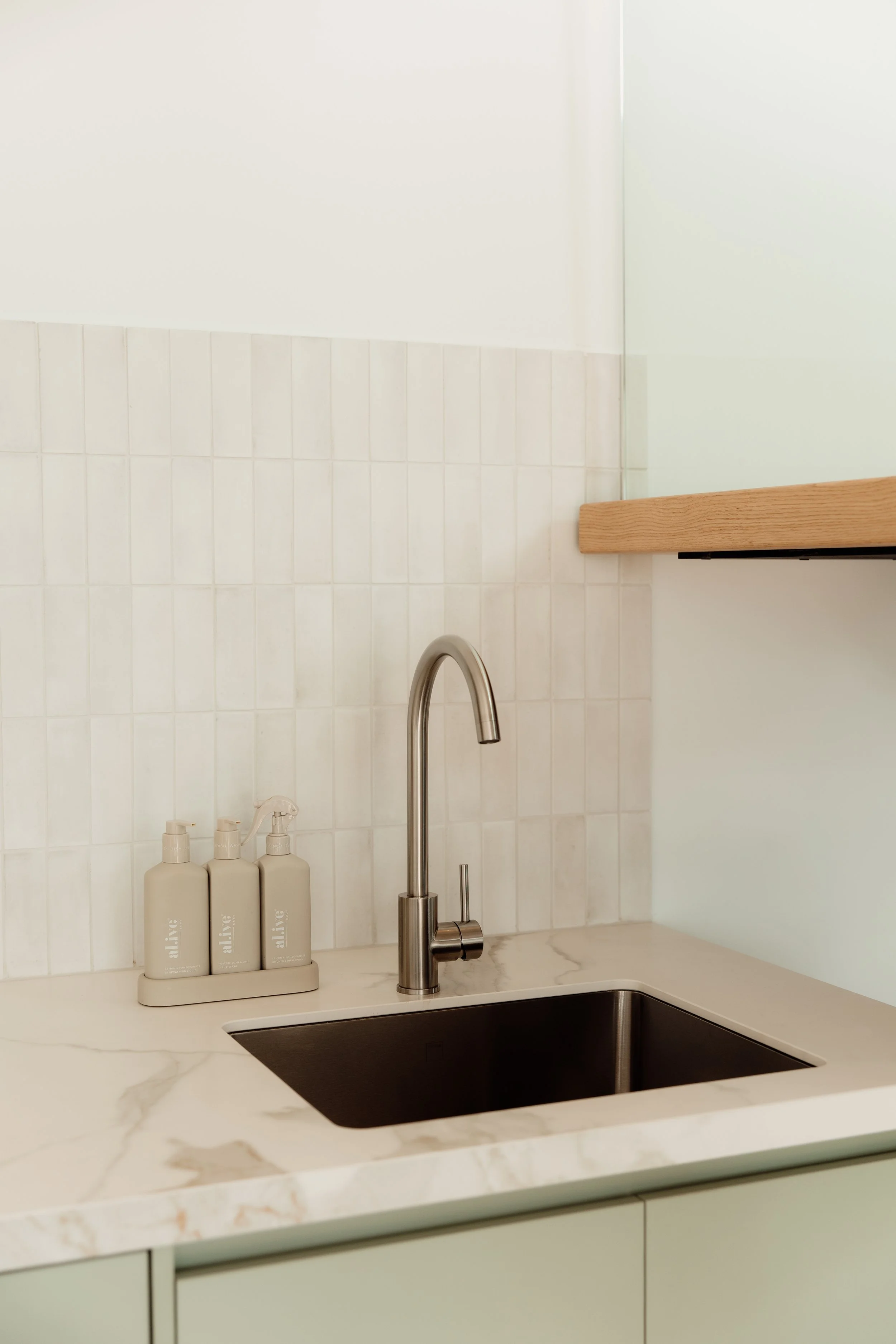 A modern kitchen sink with a stainless steel faucet, beige countertop, and bottles of skincare products labeled "alive" on a tray, with a white tiled backsplash and a wooden shelf above.