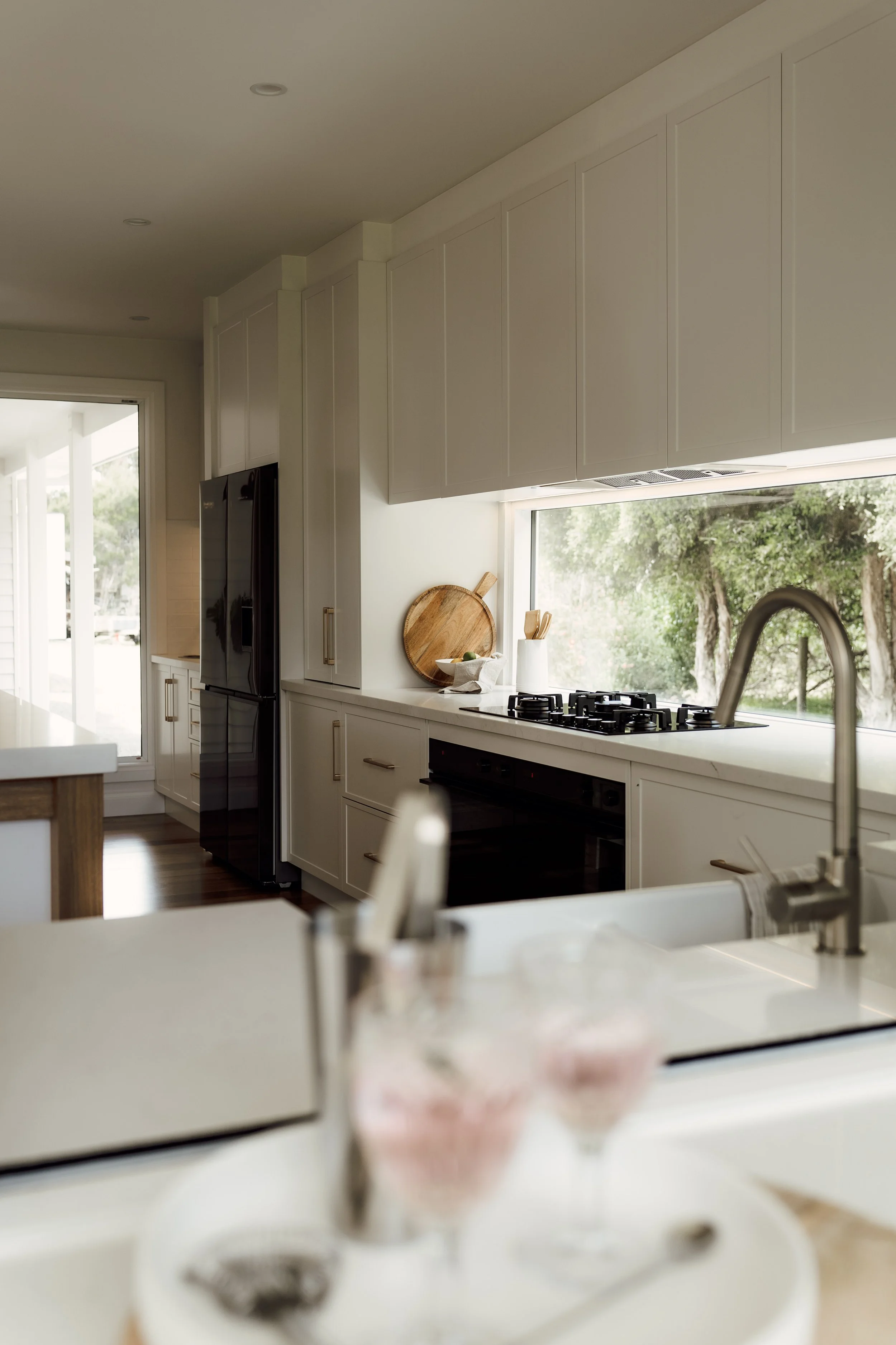 A modern kitchen with white cabinets, a black refrigerator, a window overlooking trees, a wooden cutting board, and a gas stove with a white countertop.