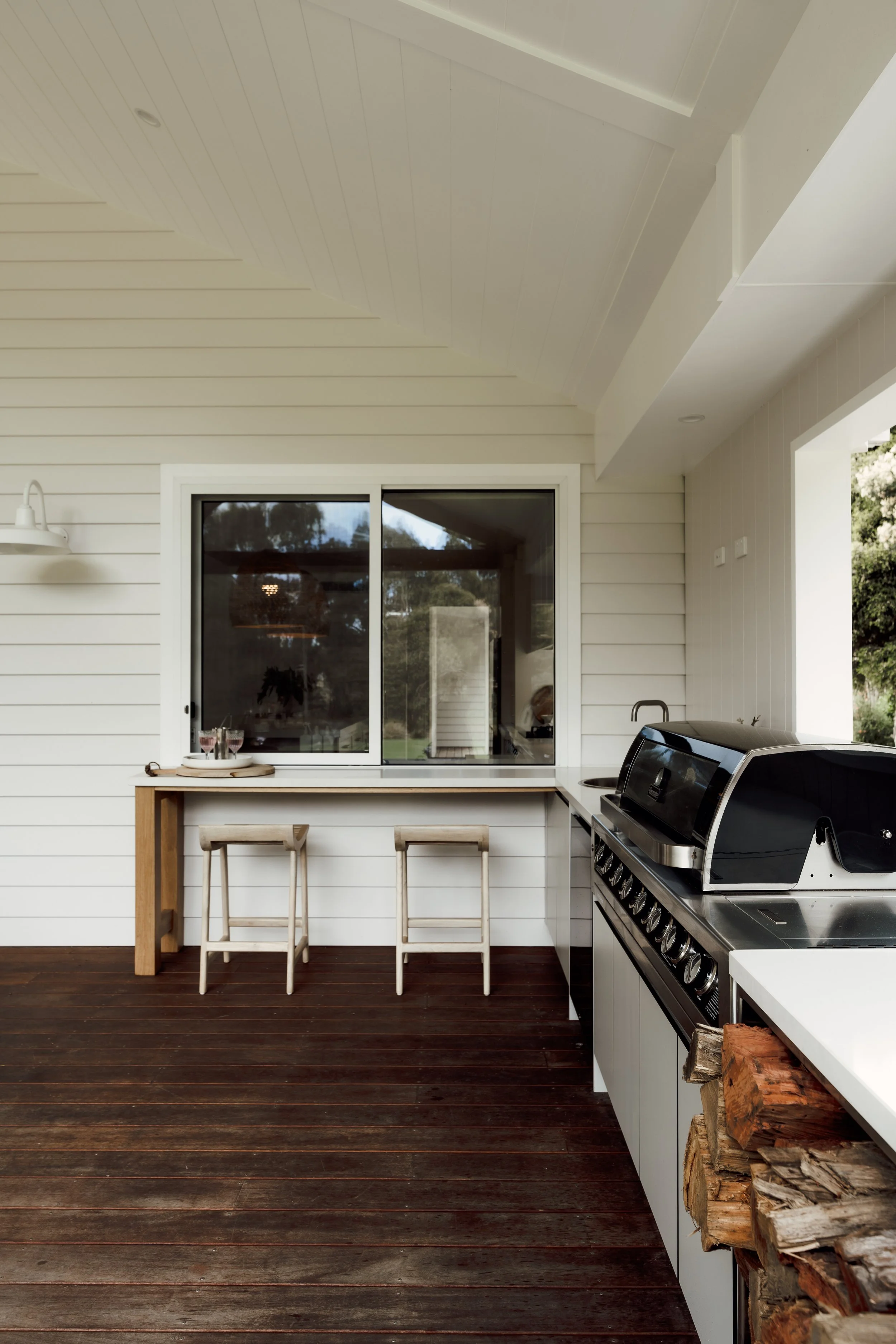 An outdoor patio kitchen with a stainless steel grill, a white countertop, a window, two wooden bar stools, and a stack of chopped firewood.
