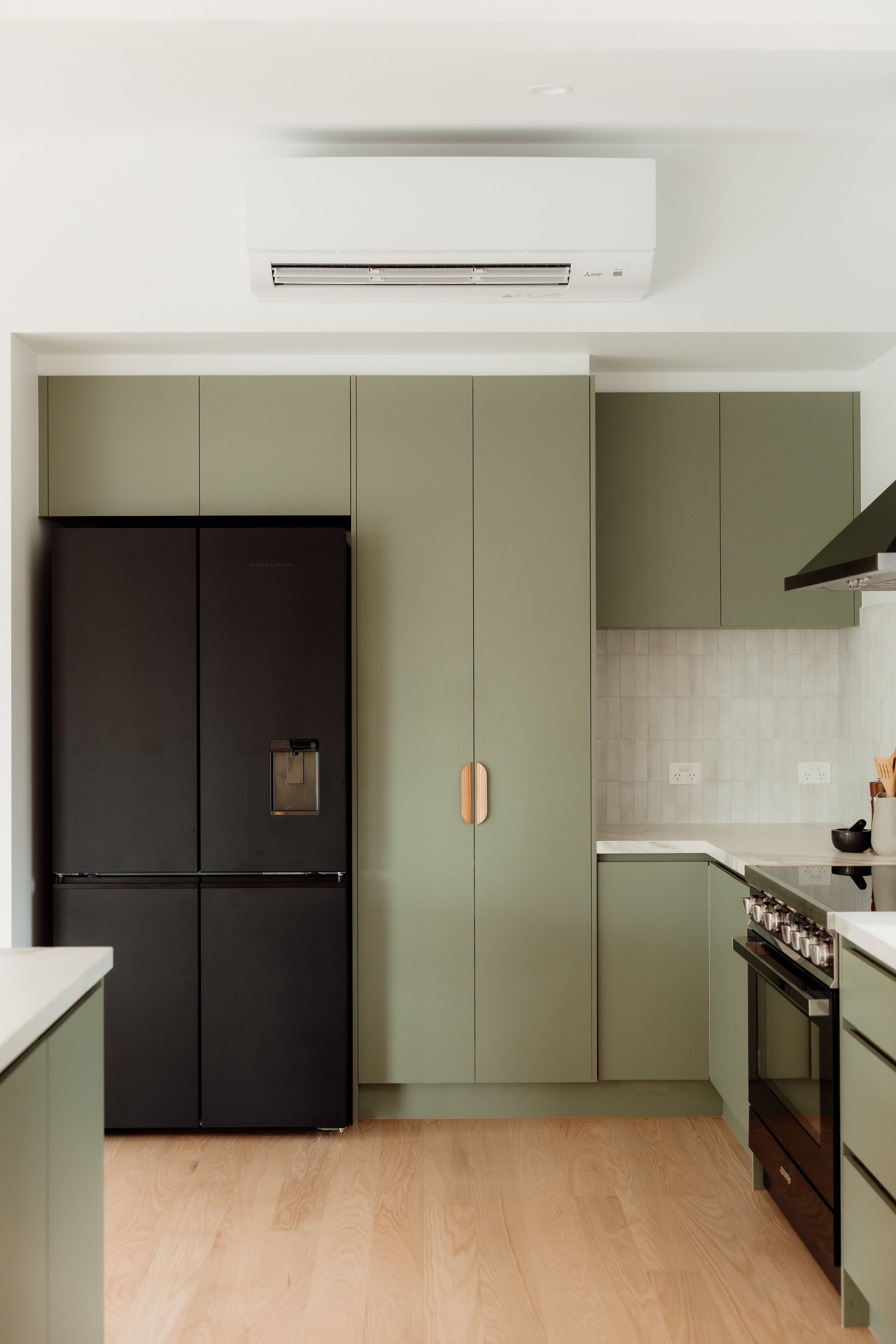 A modern kitchen with green cabinets, a black refrigerator, and a black oven, with a white countertop and light wood flooring.
