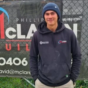 Young man wearing a blue beanie and dark hoodie, standing outdoors in front of a black fence and a promotional banner.