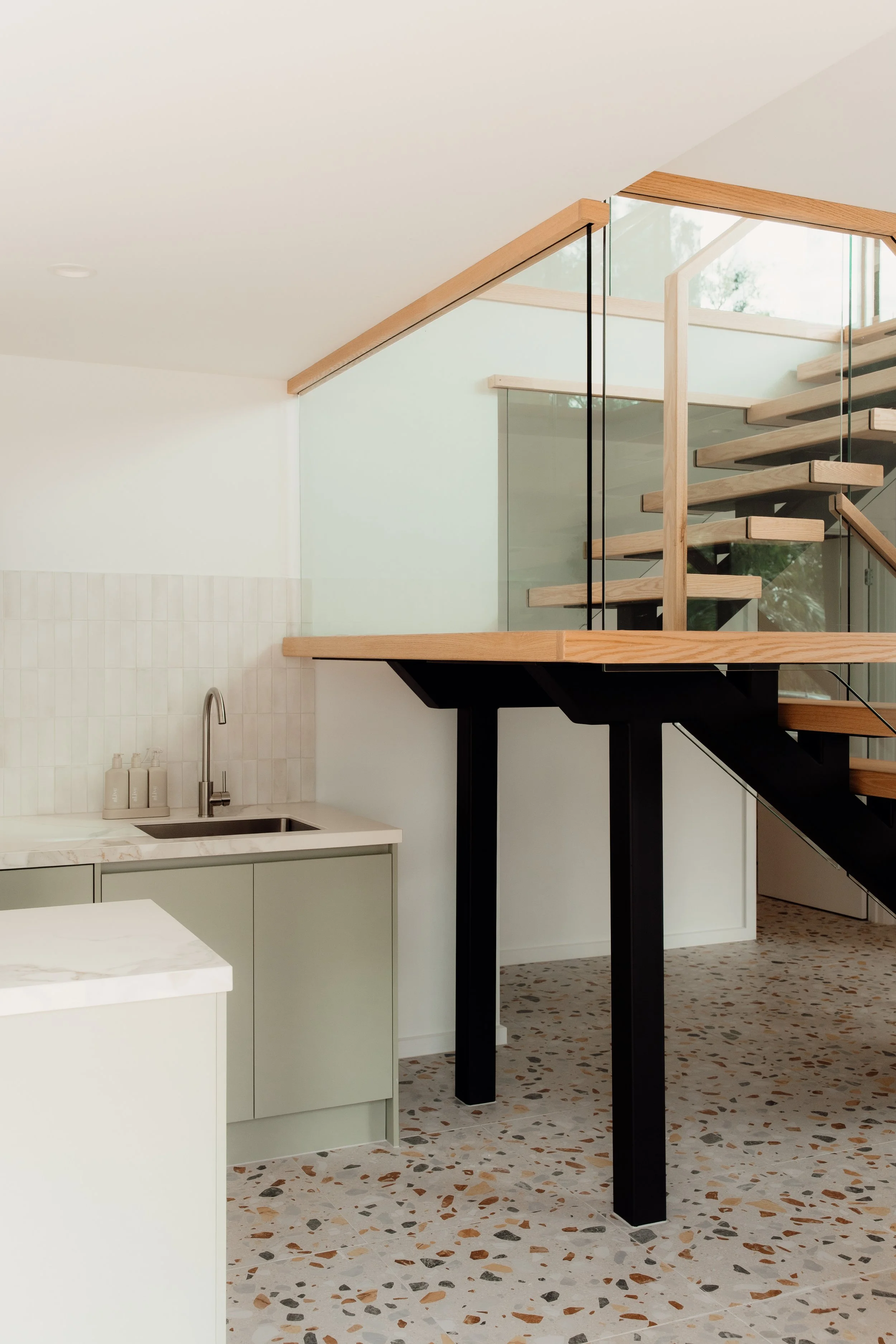 Modern interior featuring a staircase with wooden treads, a glass railing, black supports, a white kitchen with a sink and bottle containers, terrazzo flooring, and natural light coming through large windows.