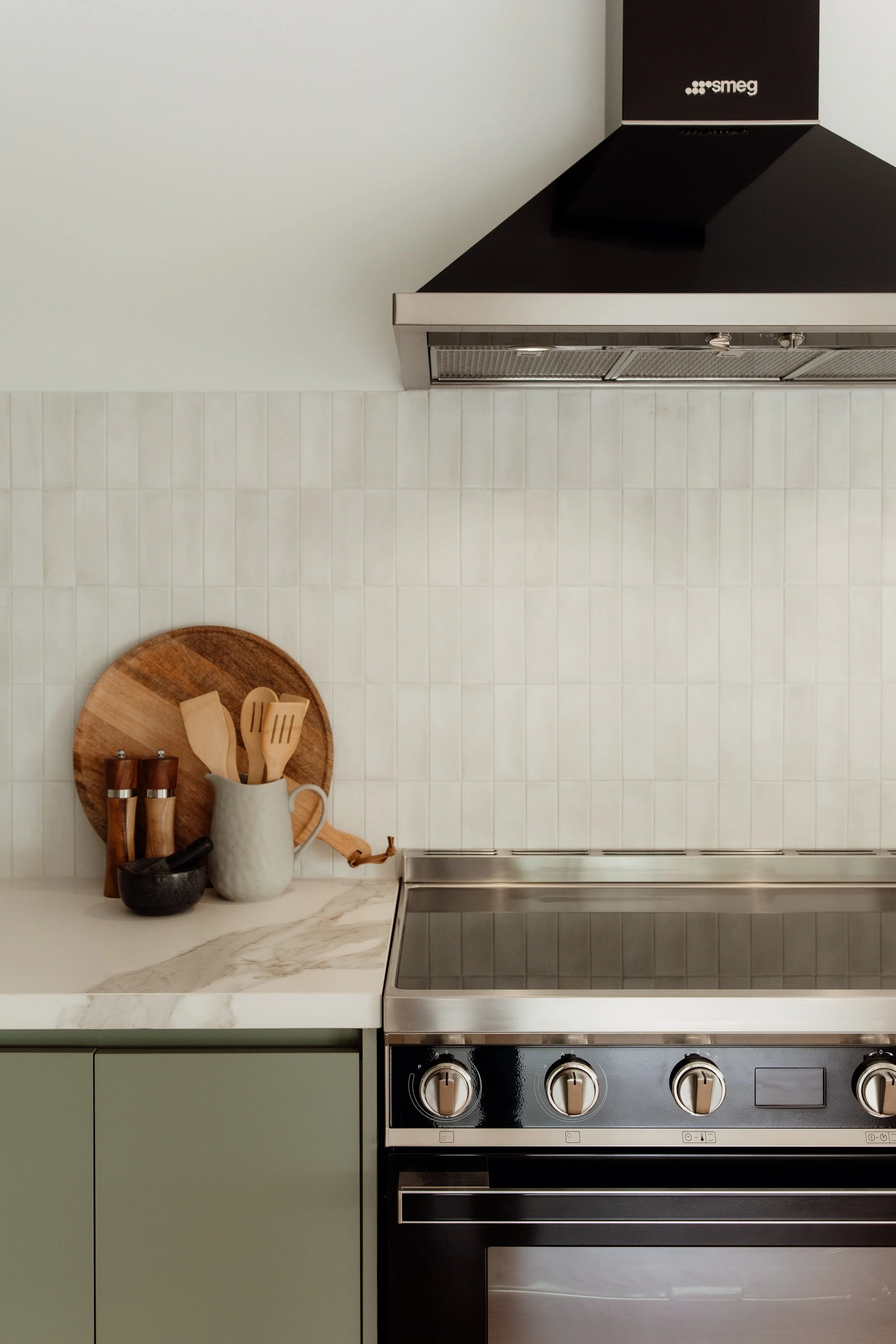 A kitchen countertop with a wooden cutting board, a white pitcher holding wooden utensils, and salt and pepper shakers. A stove with control knobs is visible below the countertop. The wall behind has white tiles and a black range hood with a silver t
