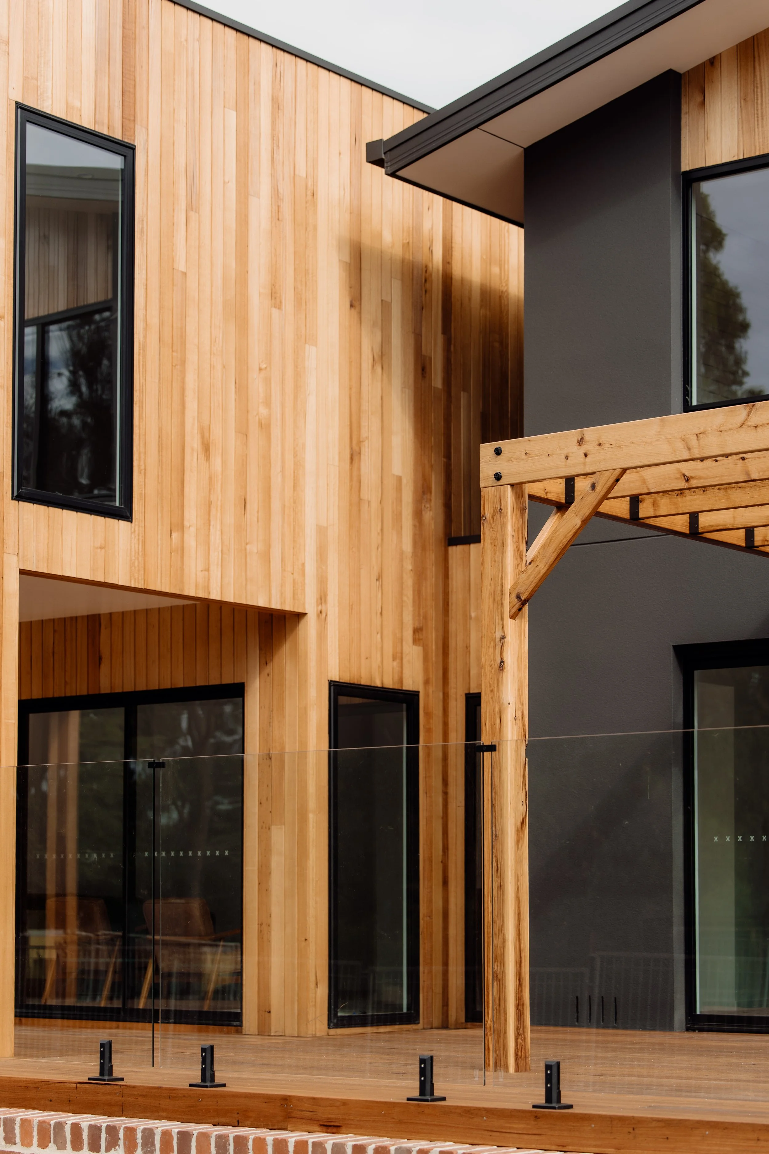 Close-up of a modern house exterior with wooden and black wall panels, large windows, and a glass balcony railing.