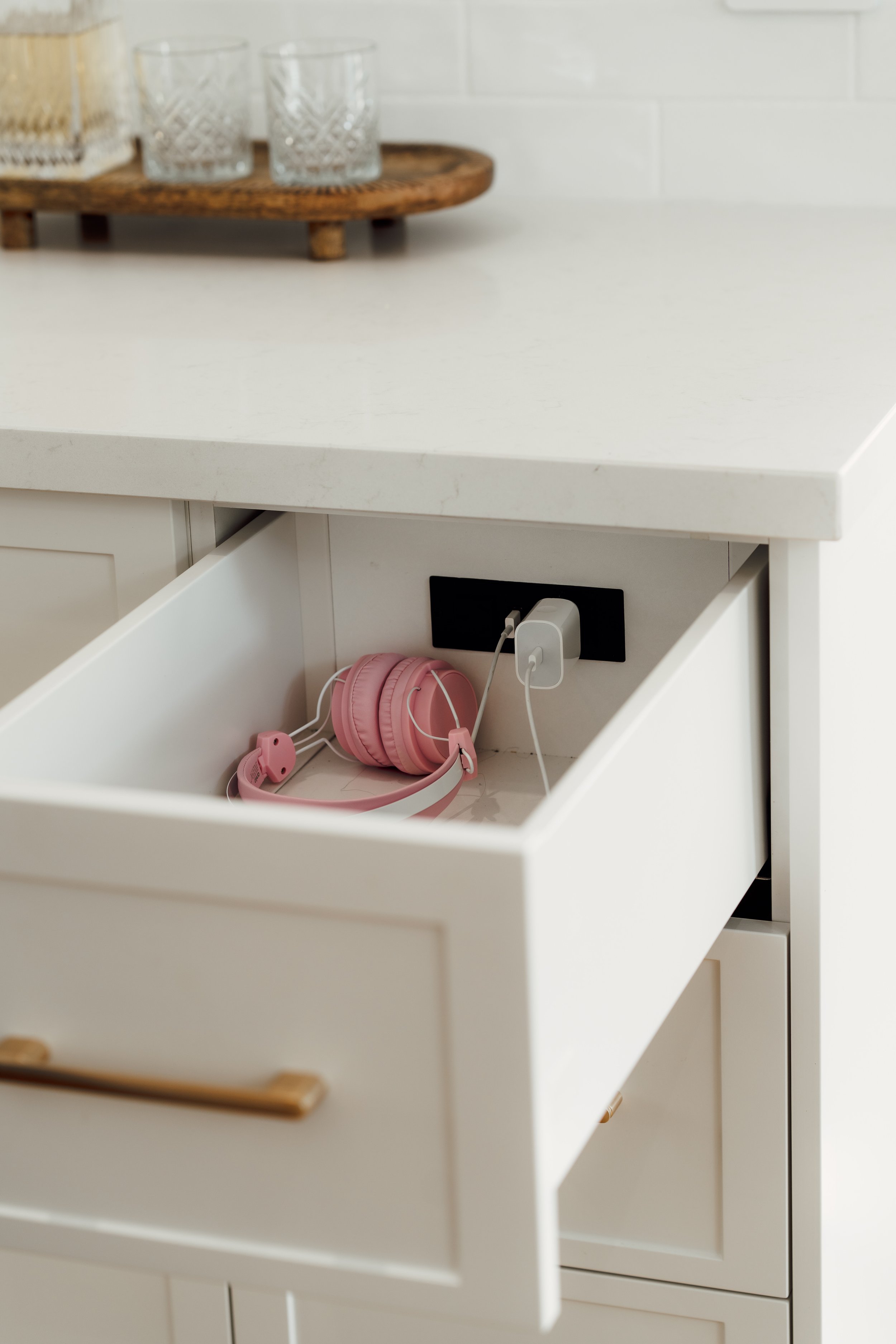 Open white drawer of a kitchen cabinet with pink headphones and a charger inside.