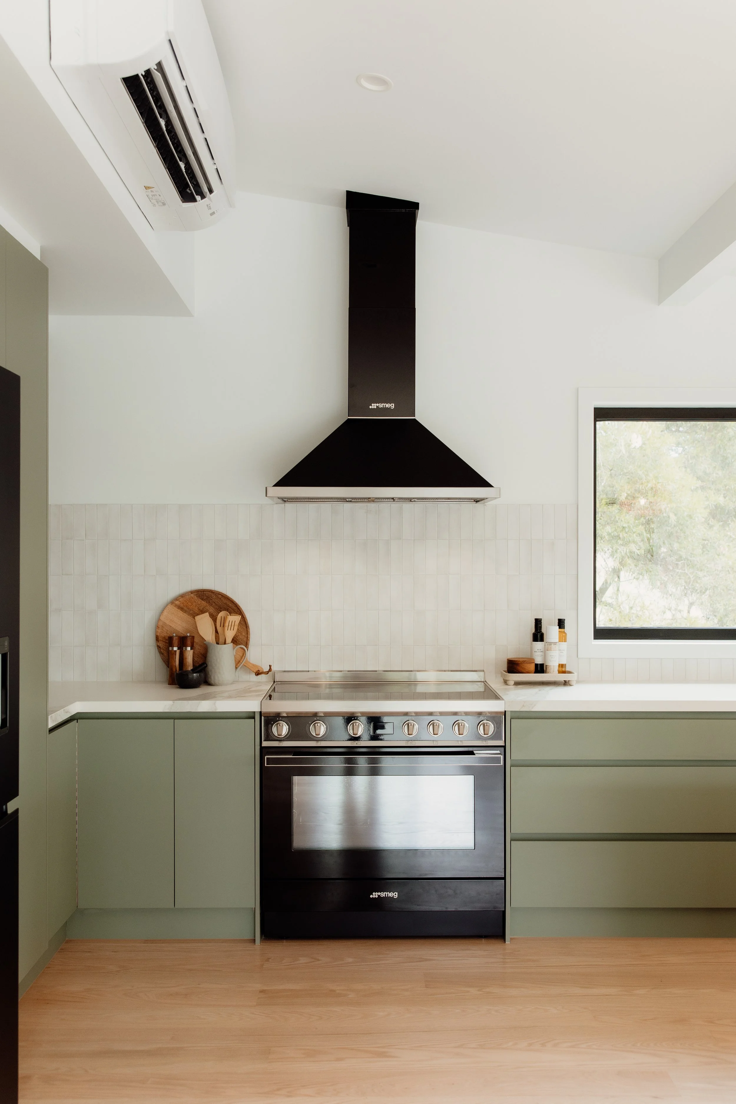 Modern kitchen with green cabinets, black stove and range hood, white tiled backsplash, window, and wooden floor.