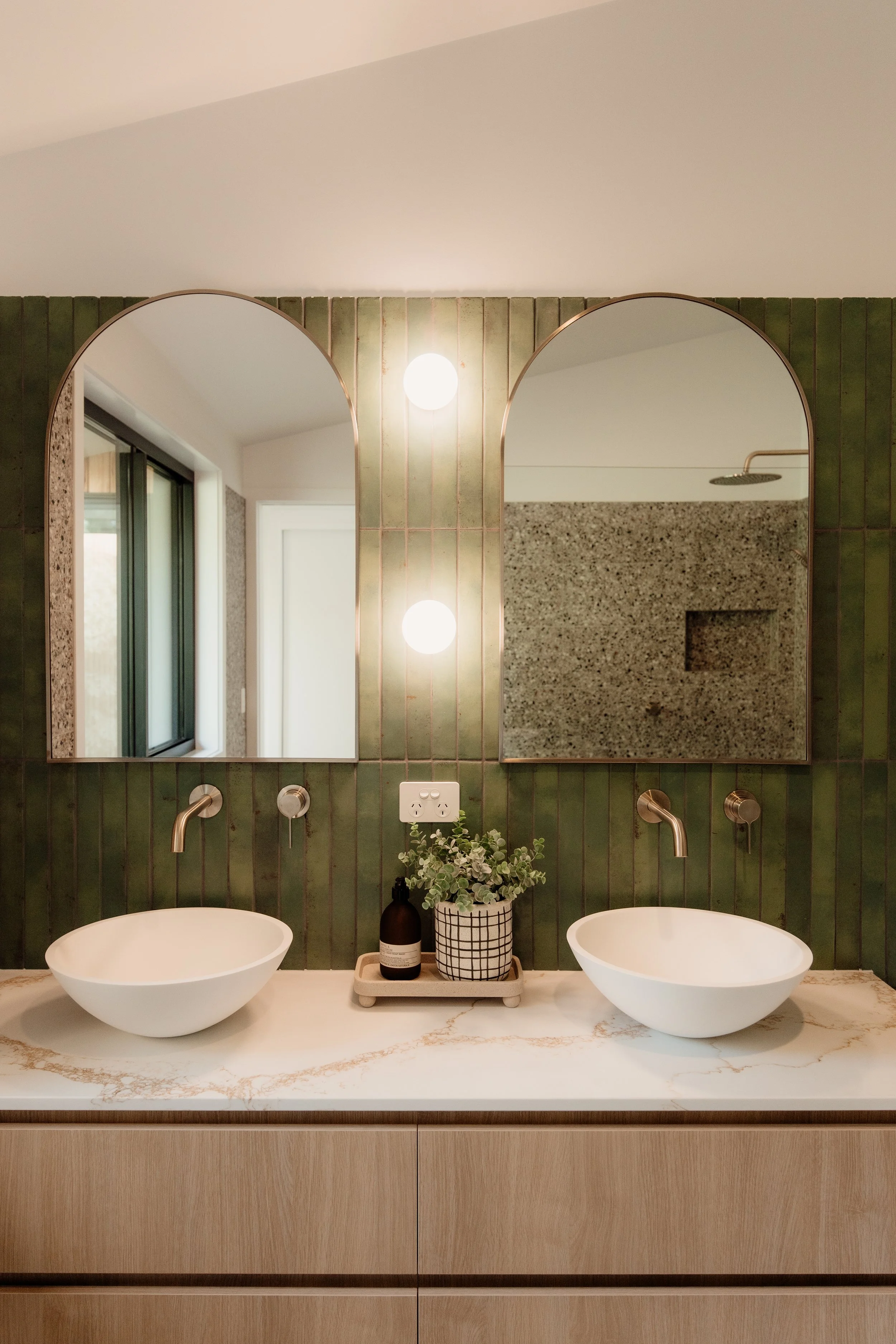Modern bathroom vanity with two vessel sinks, large mirrors, green tiled wall, pendant lights, a potted plant, and a granite shower wall.