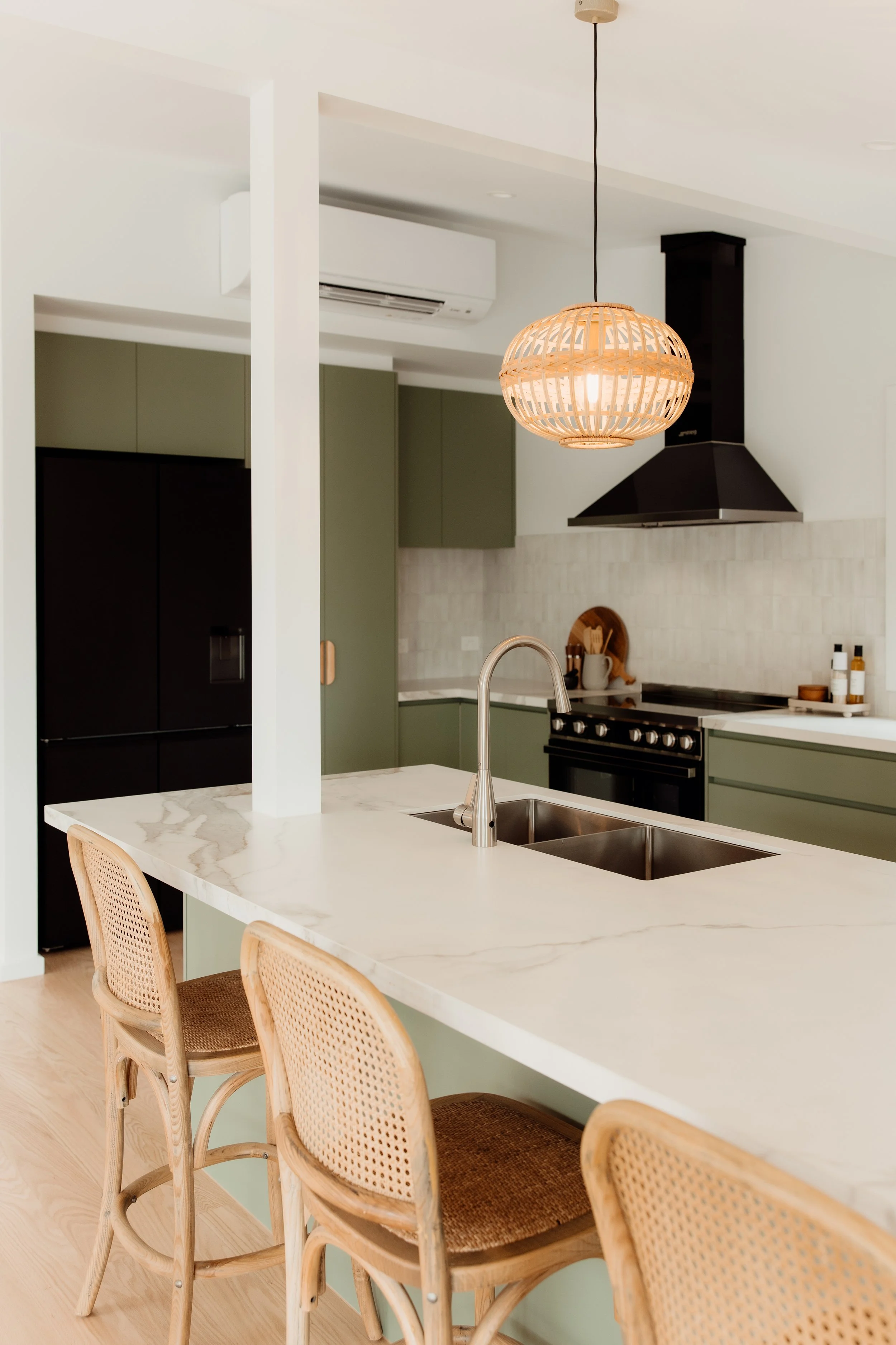 A modern kitchen with a white marble island, light wood chairs, green and black cabinetry, a black range hood, and a wicker pendant light with an exposed bulb.