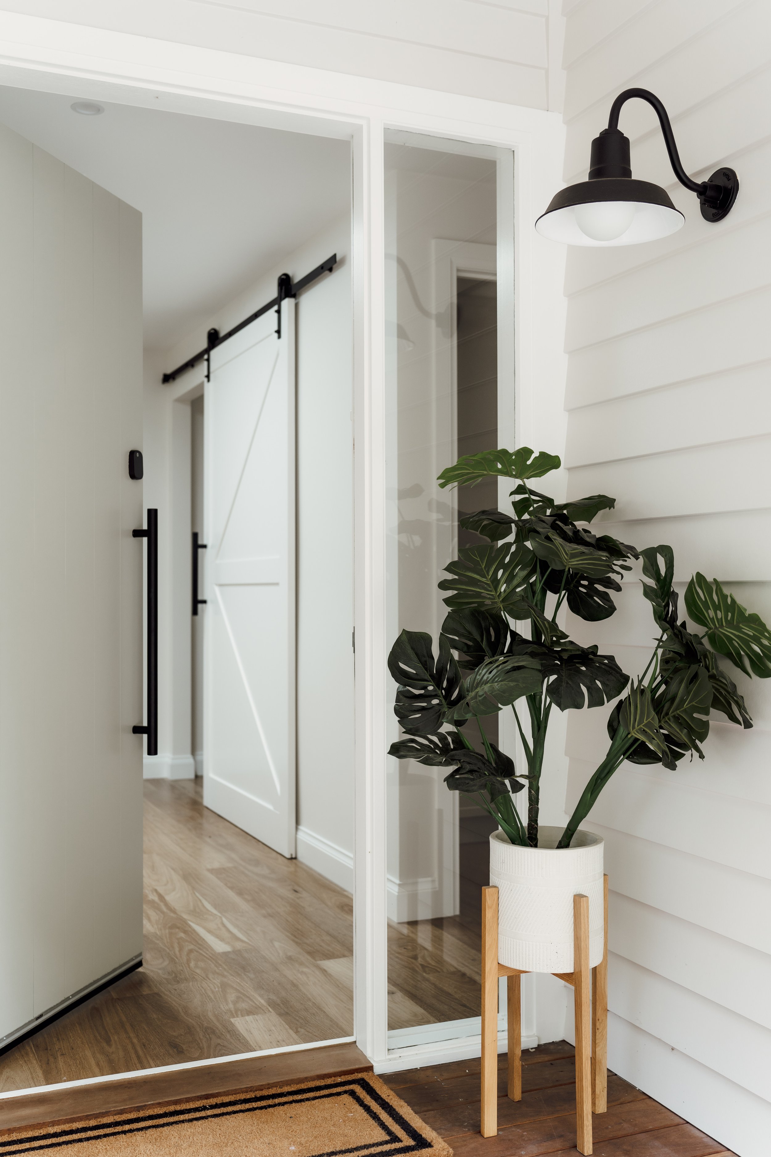 Interior of a modern home entryway featuring a plant in a white pot on a wooden stand, a white paneled wall with a black wall sconce, a large mirror, a sliding barn door, and a part of a multicolored rug on a wood floor.