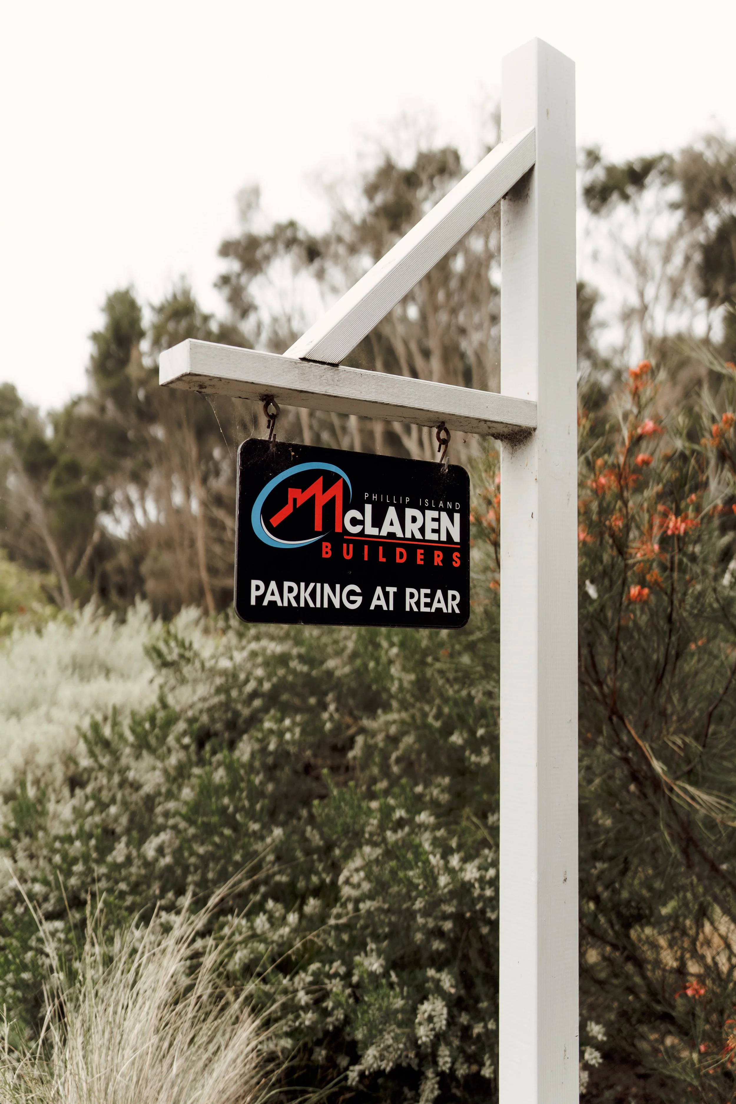 A white pole with a black and red sign that reads "Parking at Rear" and has the McLaren Builders logo, set against a background of trees and bushes.