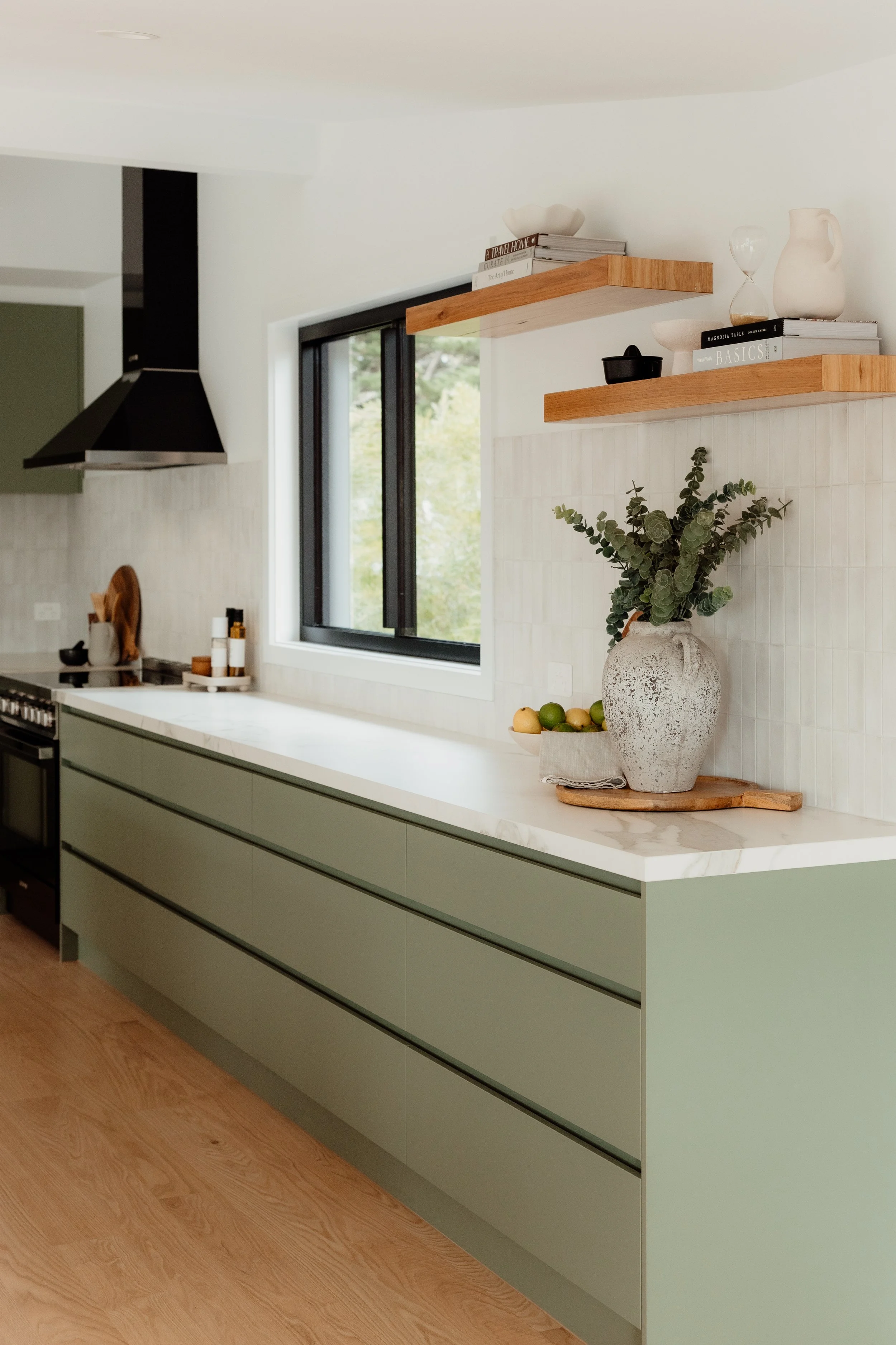 Modern kitchen with light green cabinets, white marble countertop, black range hood, and two wooden floating shelves decorated with books, vases, an hourglass, a bowl, and a black container. A large pottery vase with greenery is on the countertop wit
