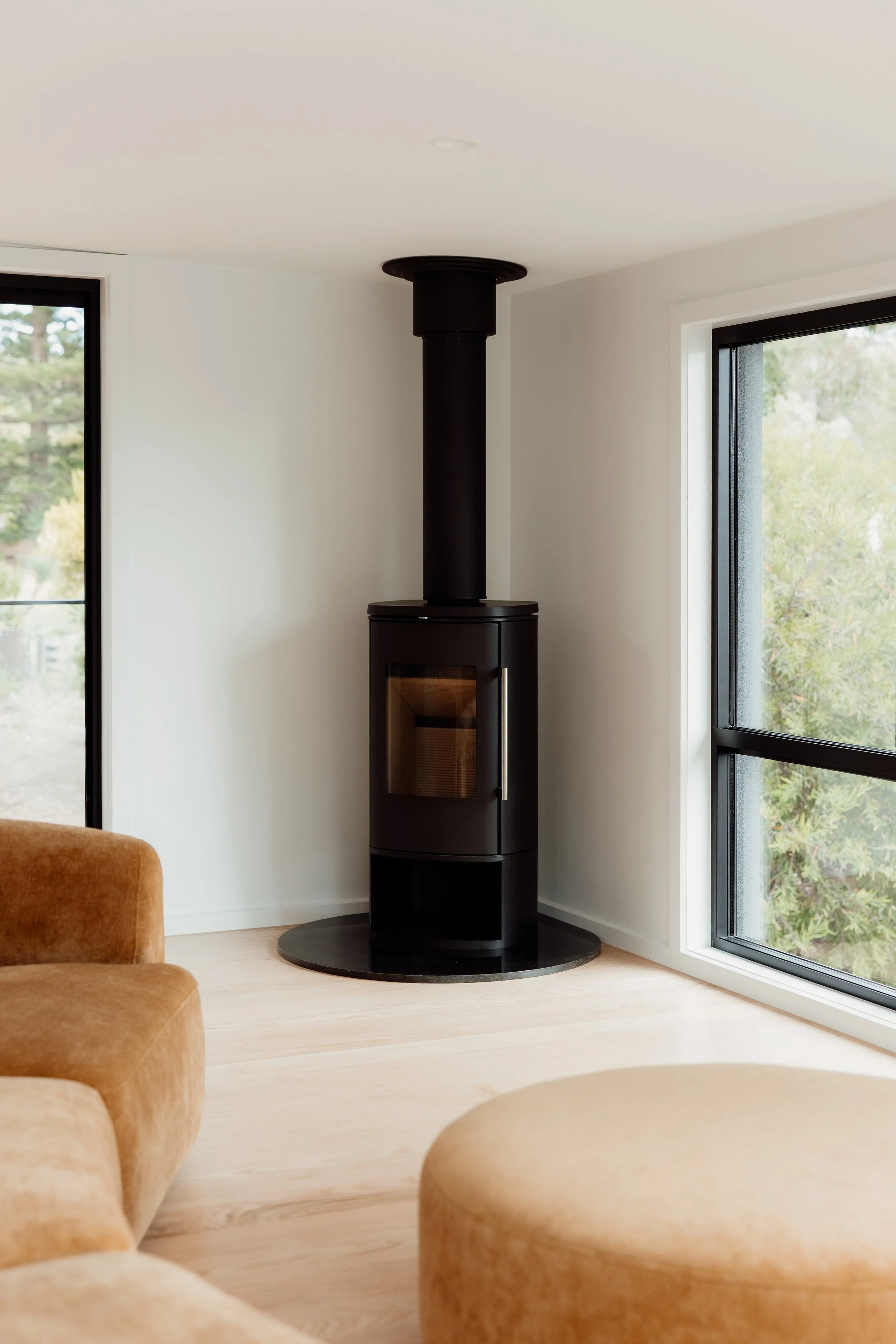 Modern living room corner with a wood stove, large windows with black frames, and tan seating.