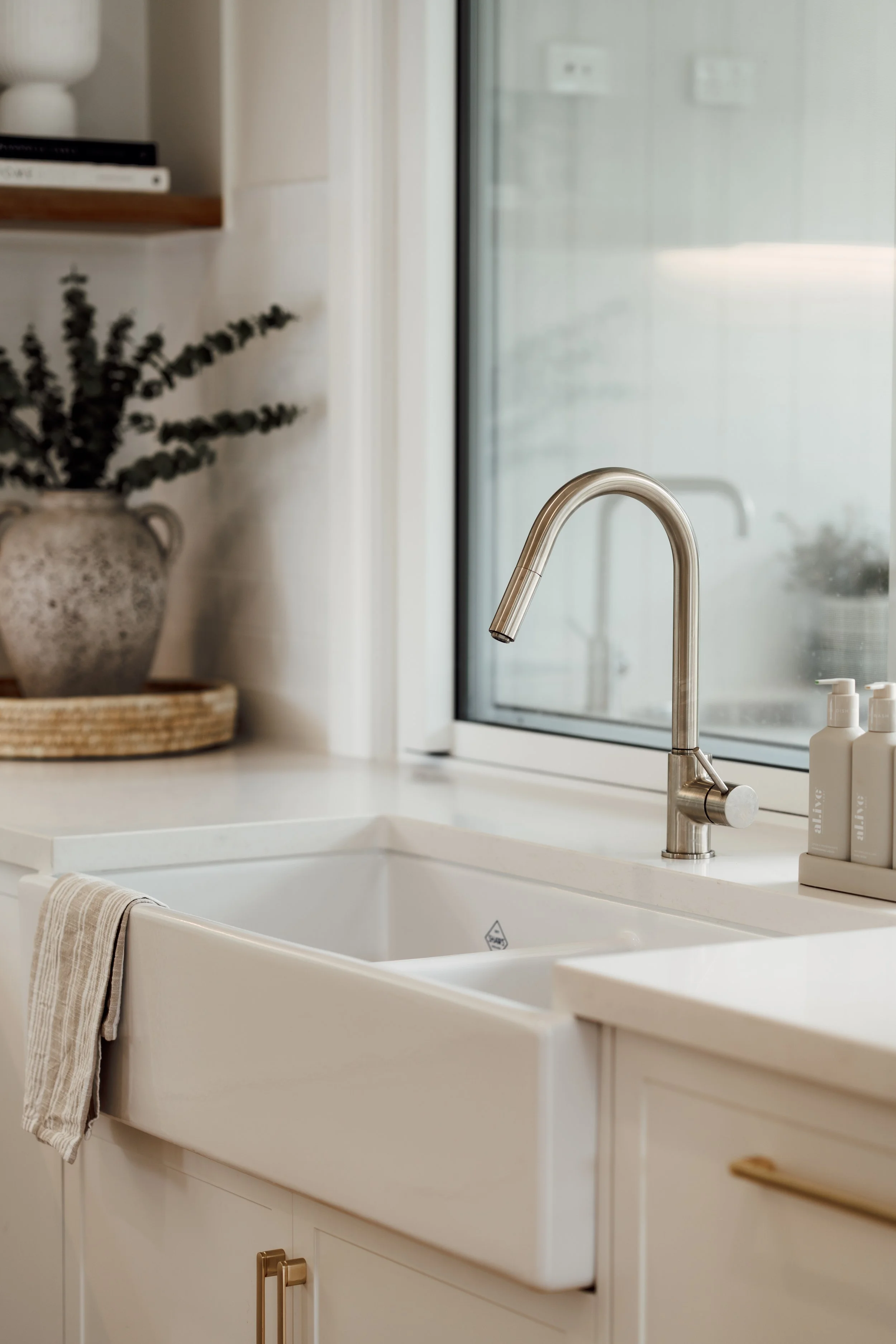 Modern kitchen with white sink, metallic faucet, soap dispensers, and a window bringing natural light, with a potted plant and books on a shelf in the background.