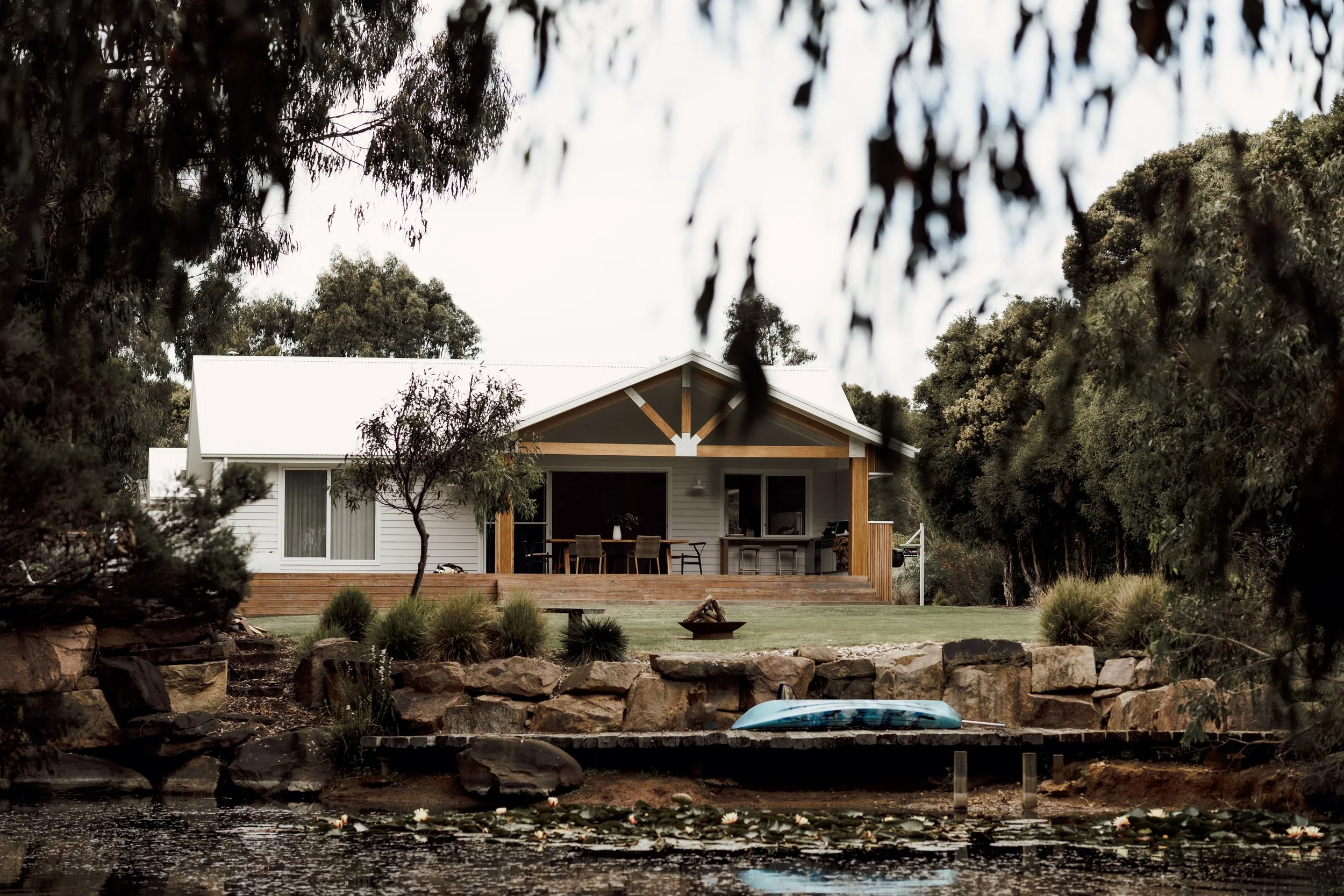A house with a white exterior and a deck, surrounded by trees and shrubs, with a pond or small lake in the foreground featuring floating lily pads and a blue kayak.